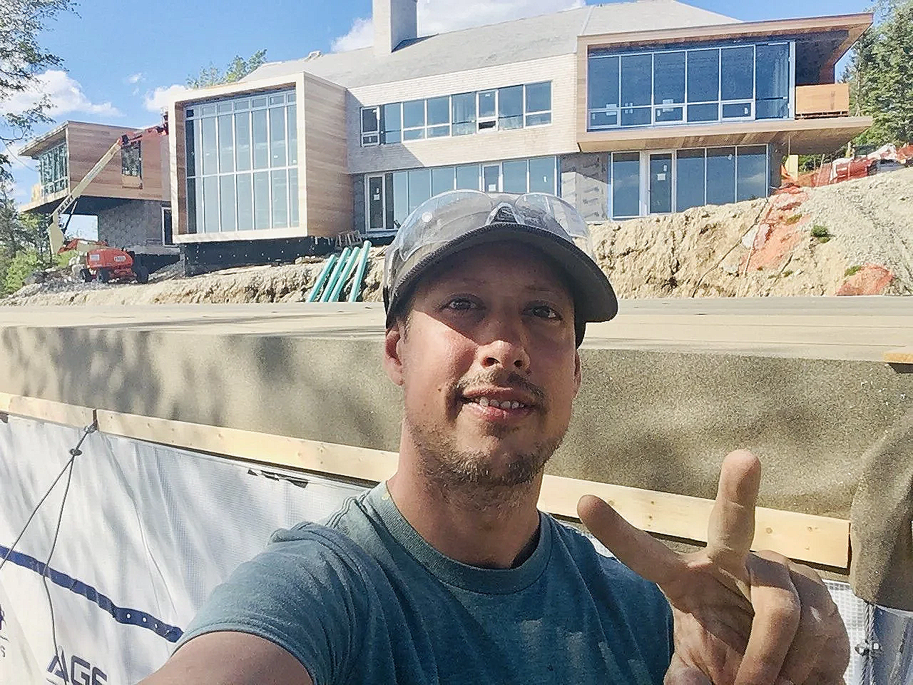 A man taking a selfie at a construction site with a house under construction in the background. The man is wearing a cap and making a peace sign.