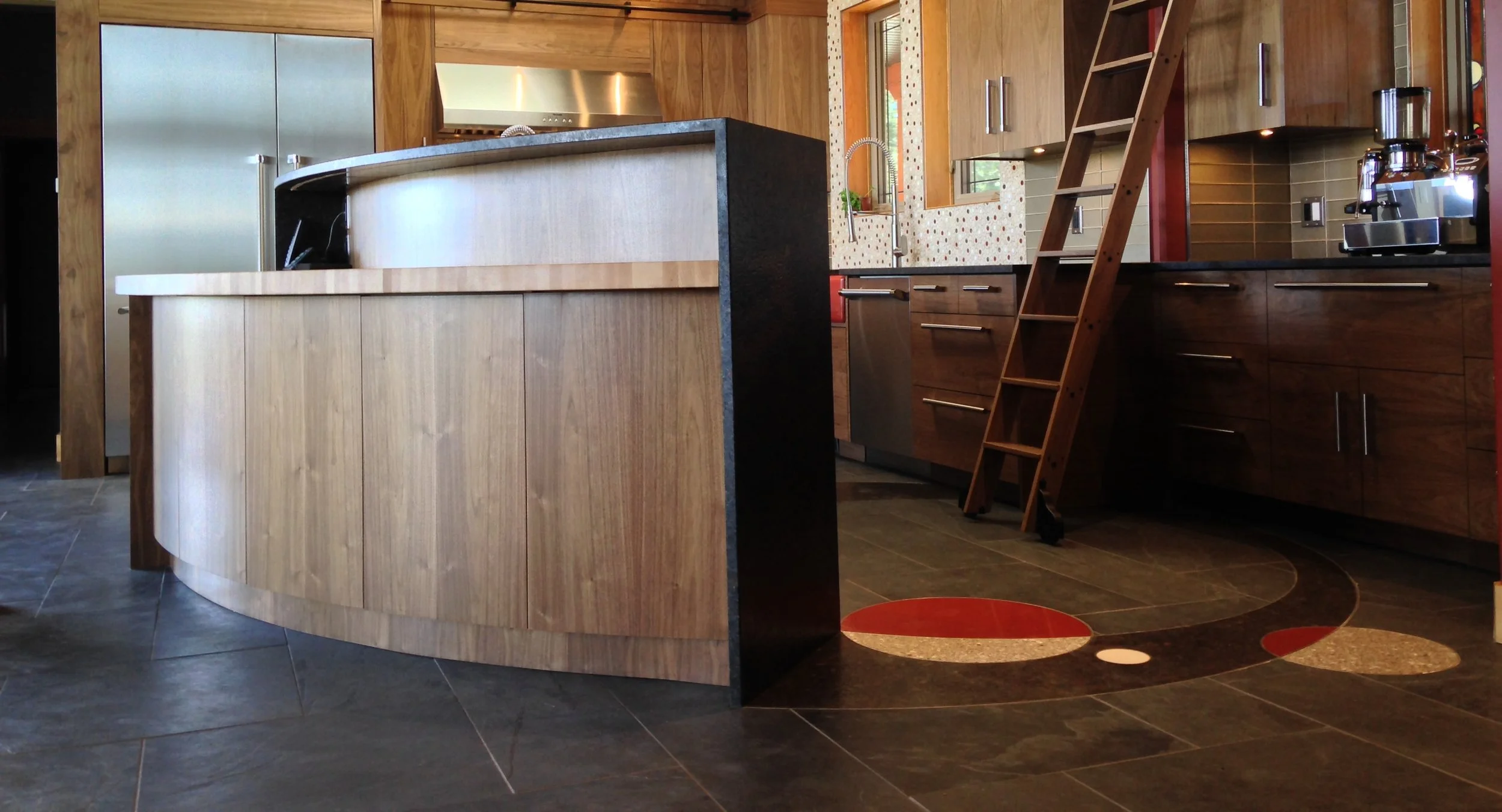 Modern kitchen with wooden cabinets, a curved island, a ladder, and a patterned backsplash, with dark tiled flooring.