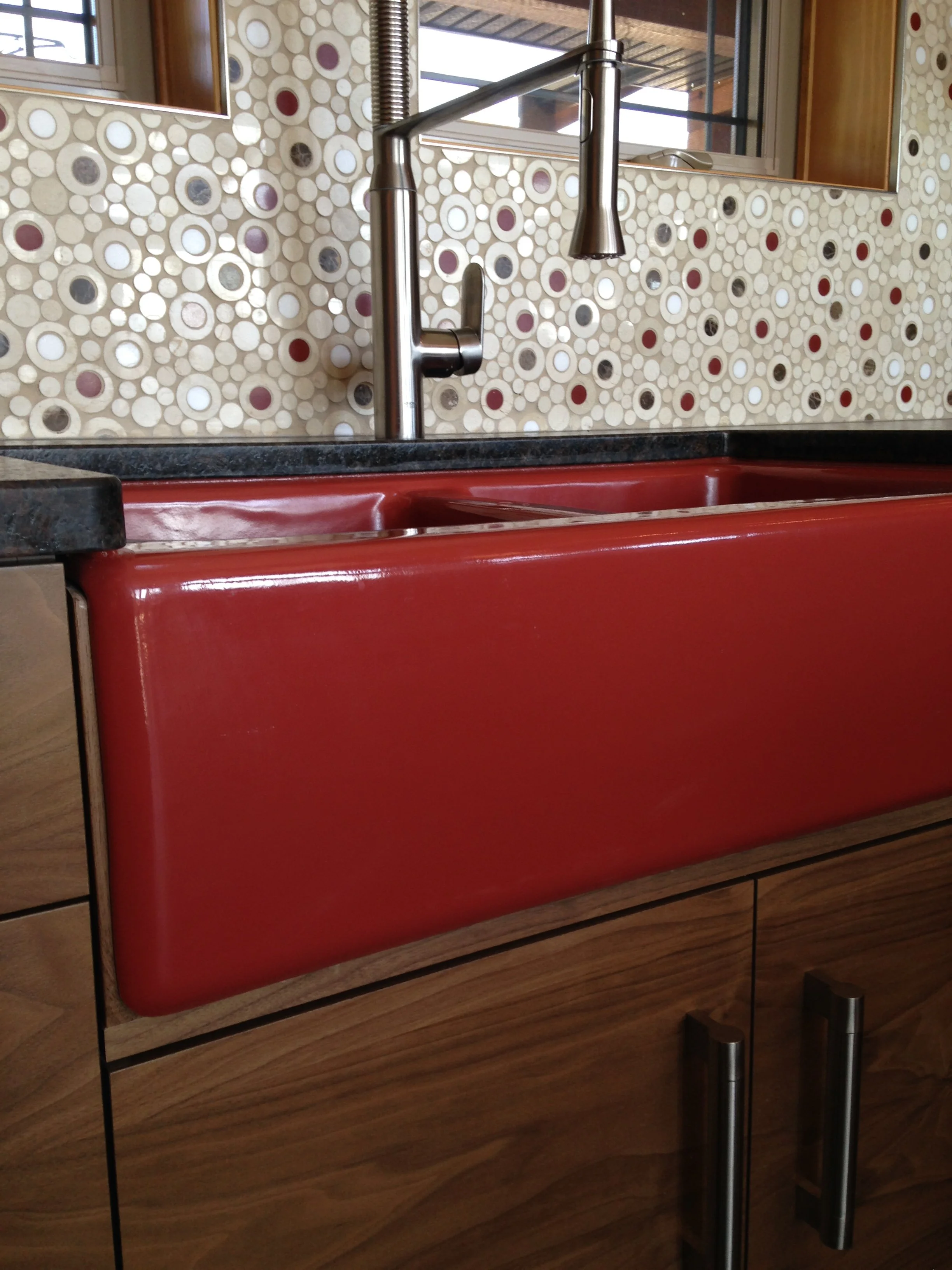 Close-up of a red farmhouse sink with wooden cabinetry below and a black countertop above. A stainless steel faucet is mounted on the countertop. The background features a tile backsplash with a circular pattern of various colors, and a window above.