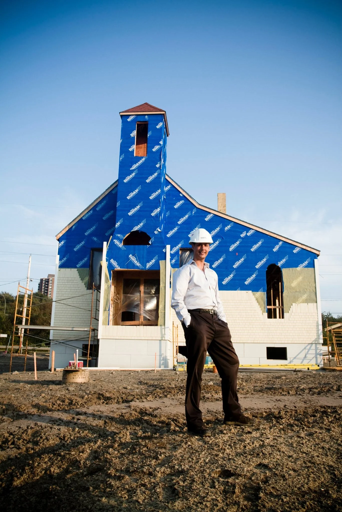A man in formal attire and a hard hat standing outdoors at a construction site with a house under construction in the background.