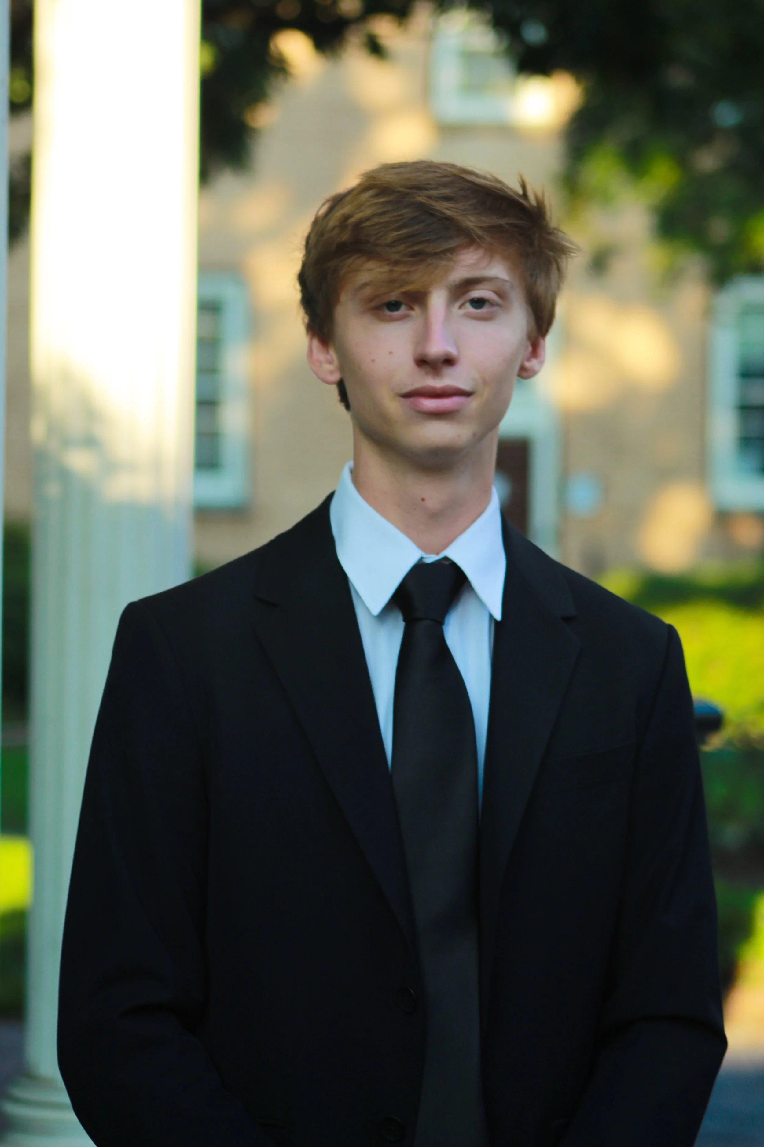 A young man with light skin and light brown hair styled to the side, dressed in a black suit, white shirt, and black tie, standing outdoors in front of a building with greenery and trees in the background.
