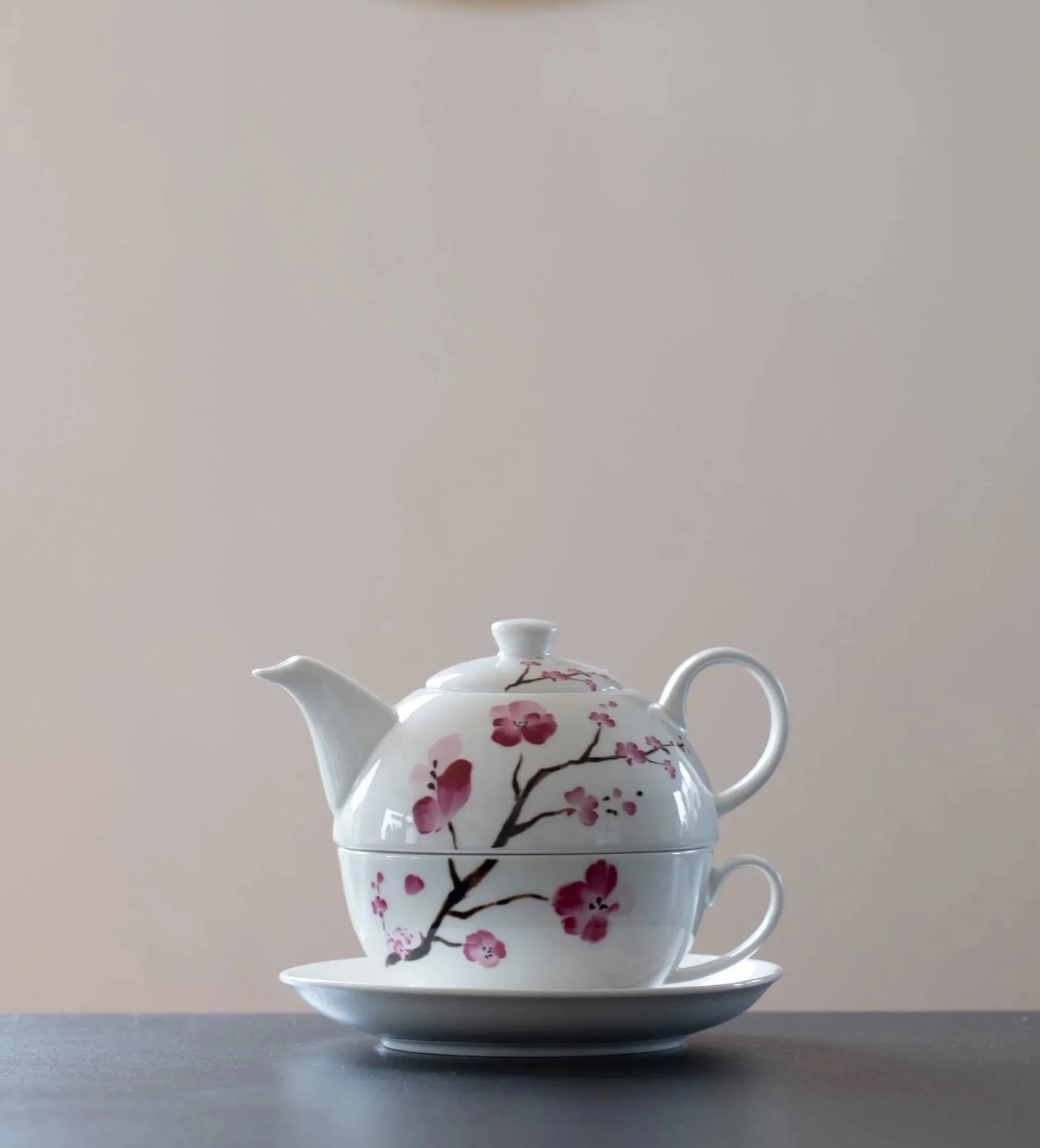 Weiße Teekanne mit Kirschblütenmuster, auf einem kleinen weißen Teller, vor einem neutralen Hintergrund.