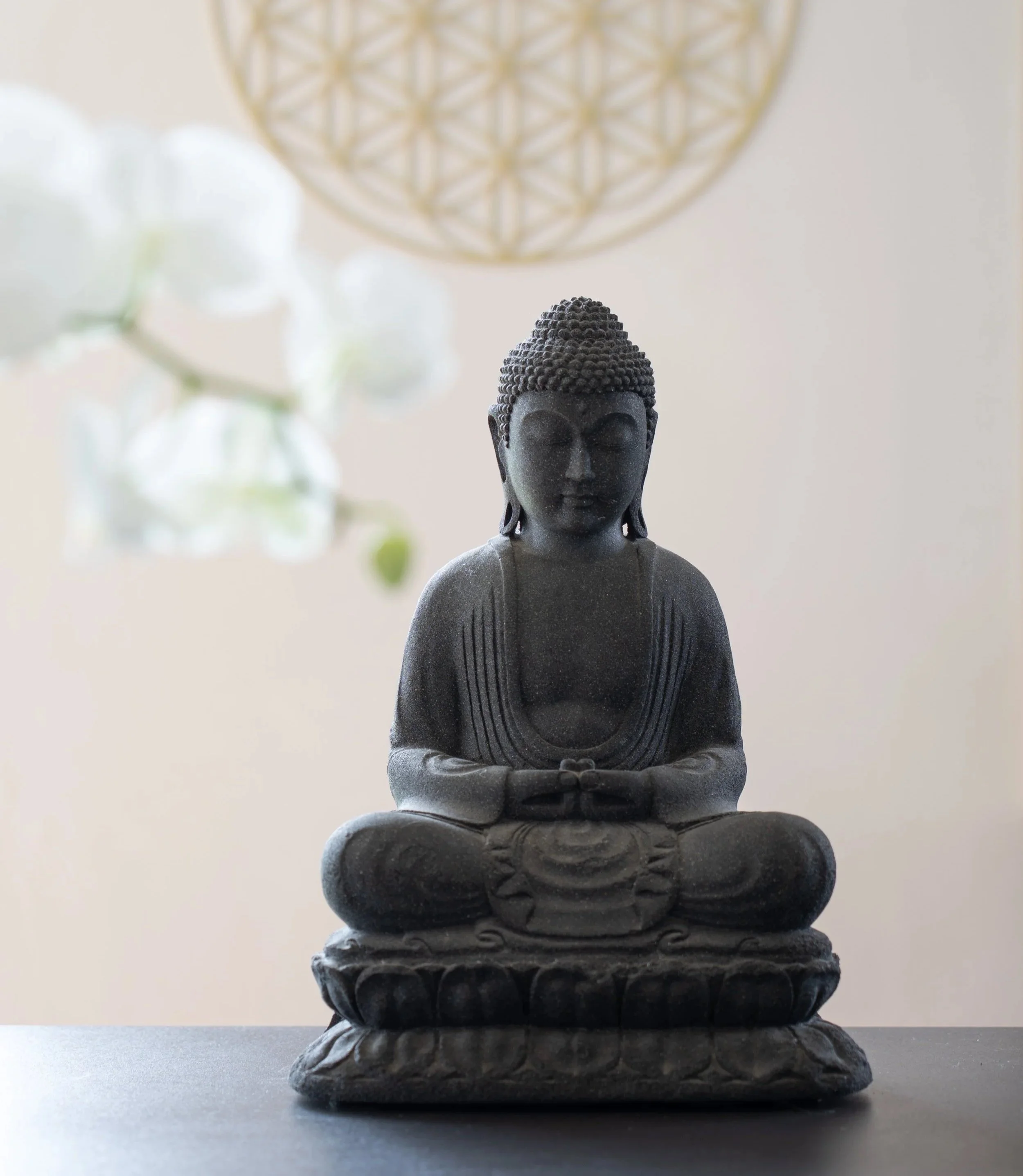 Eine schwarze Buddha-Statue in Meditationspose auf einem schwarzen Sockel, im Hintergrund eine weiße Wand und eine weiße Orchidee.