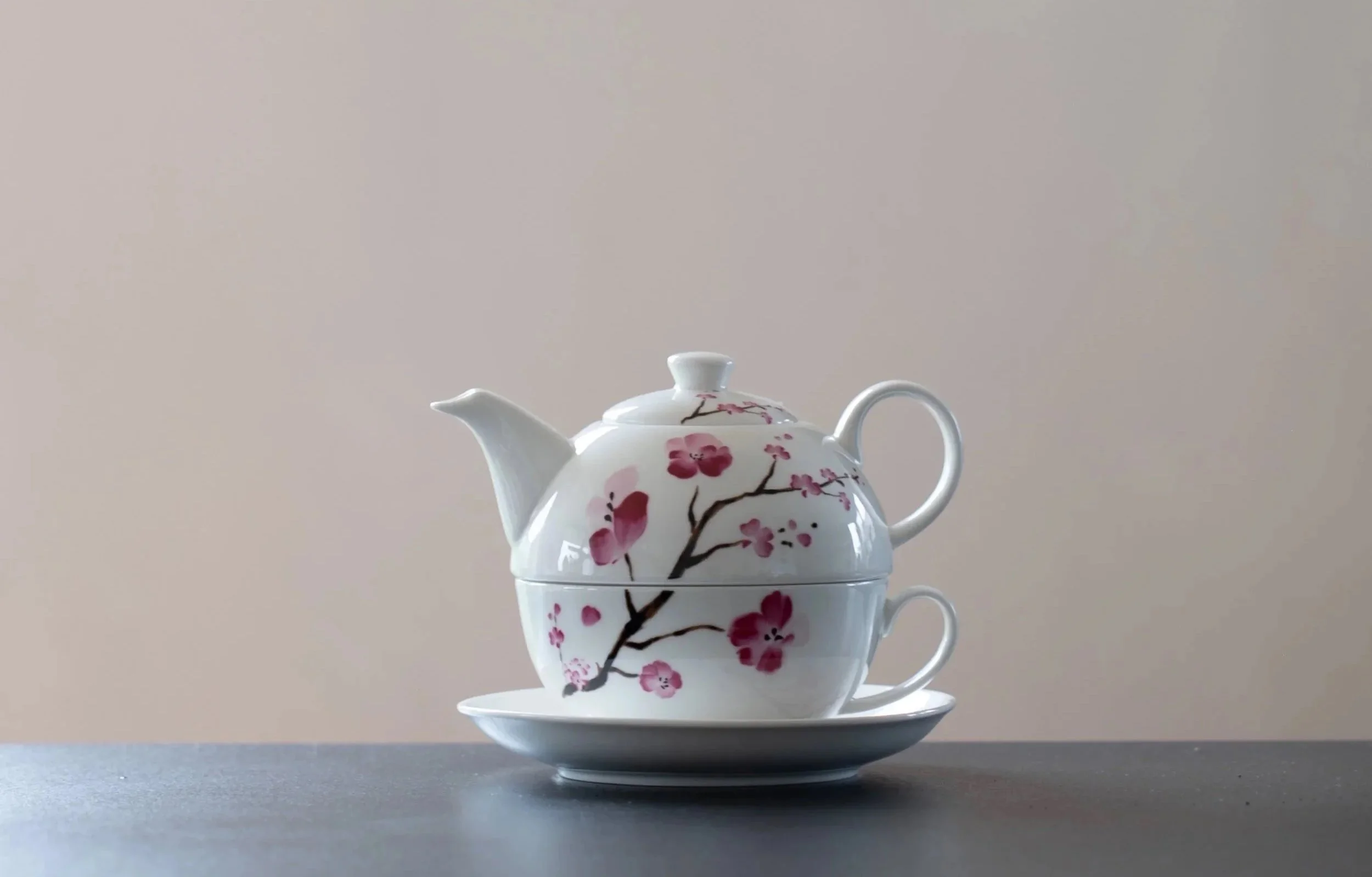 Weiße Teekanne mit rosa Blütenmuster, auf einem weißen Teller, vor einem hellen Hintergrund.