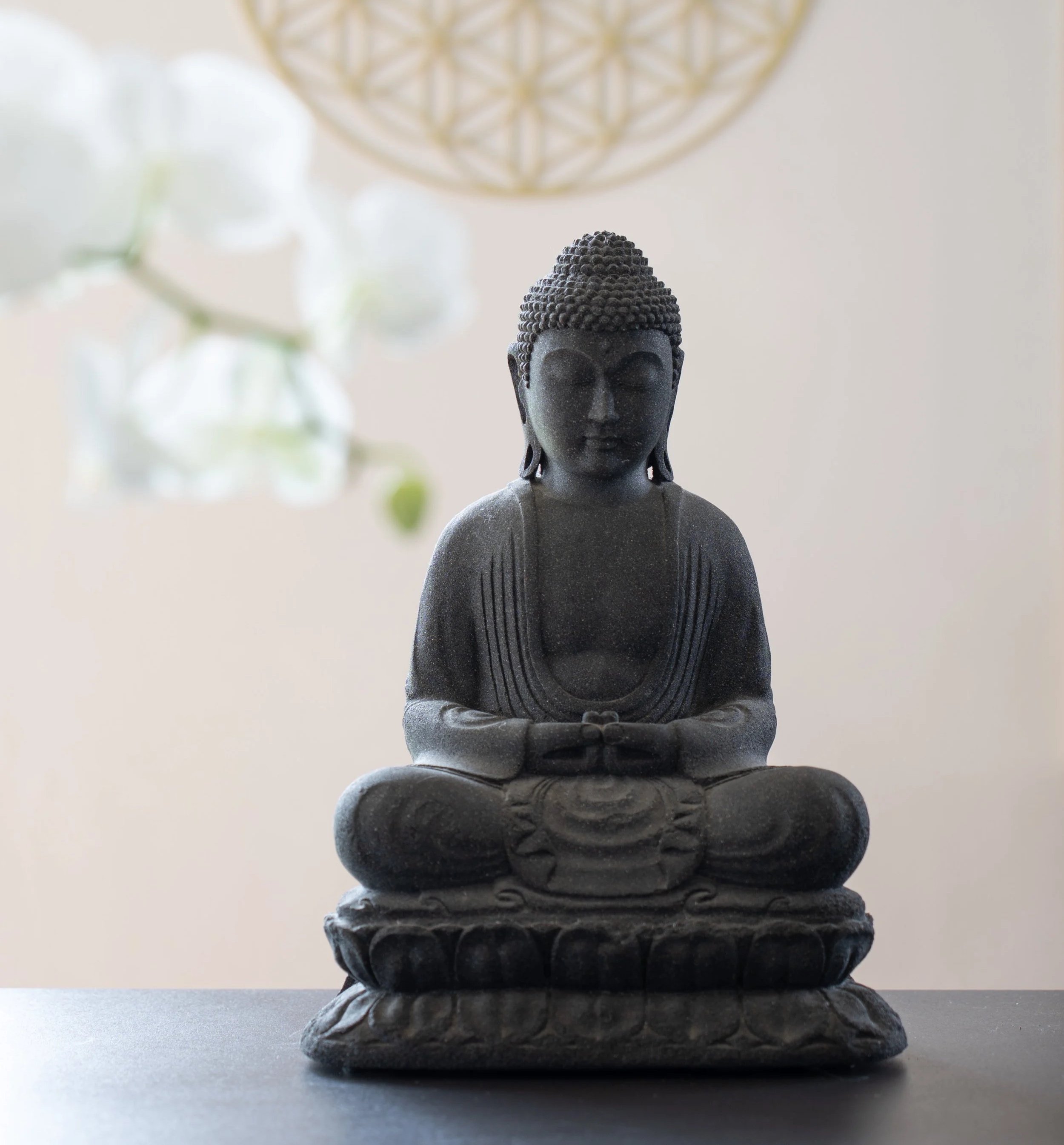 Schwarze Buddha-Figur sitzt im Schneidersitz, auf einer lotusförmigen Basis, vor einem weißen Hintergrund mit einer Blume und einem dekorativen Element an der Decke.