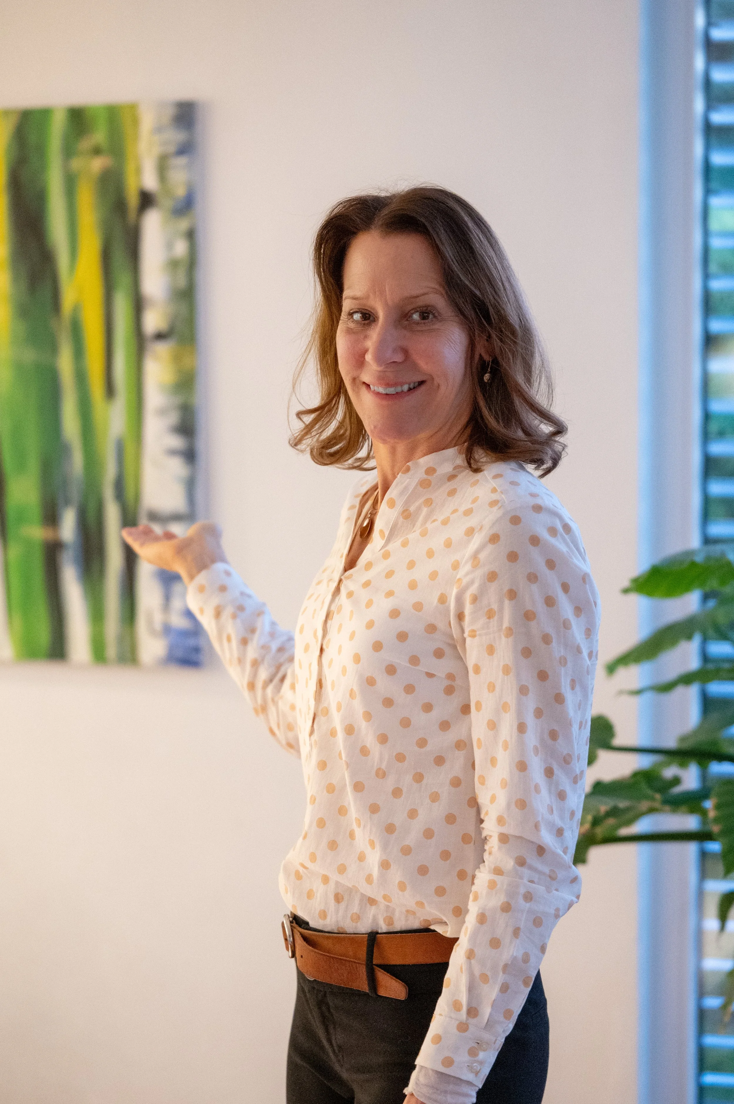 Eine lächelnde Frau in einem weißen Poloshirt mit gelben Punkten, die auf ein abstraktes Gemälde in einem Raum zeigt.