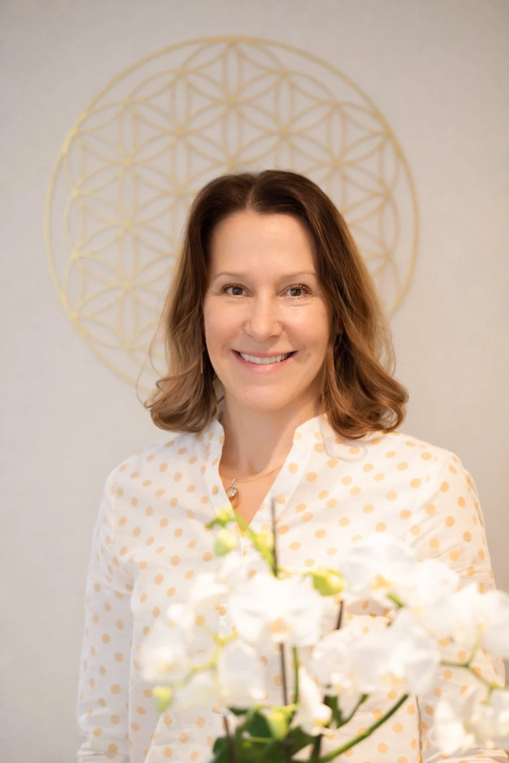 Eine lächelnde Frau mit braunen, lockigen Haaren, die ein weißes Hemd mit orangen Punkten trägt, steht vor einer goldenen geometrischen Wanddekoration und hält weiße Orchideen in der Hand.