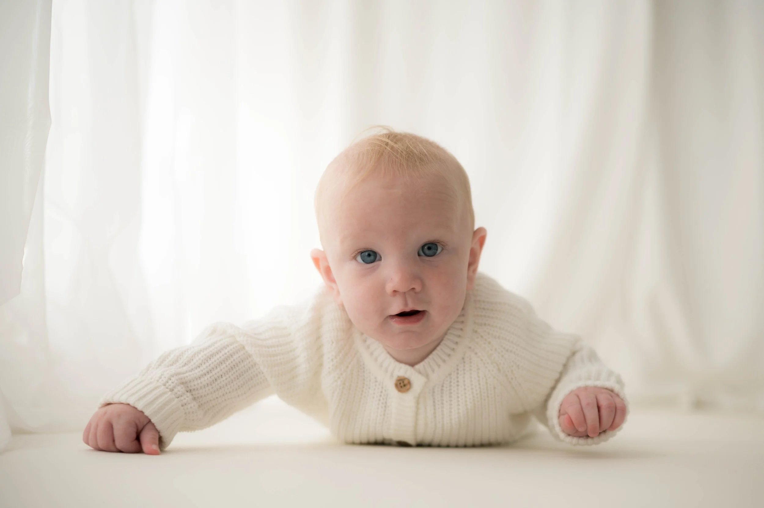 A baby with blue eyes and light hair, wearing a cream-colored knitted sweater, is crawling on a white surface with a soft white background.
