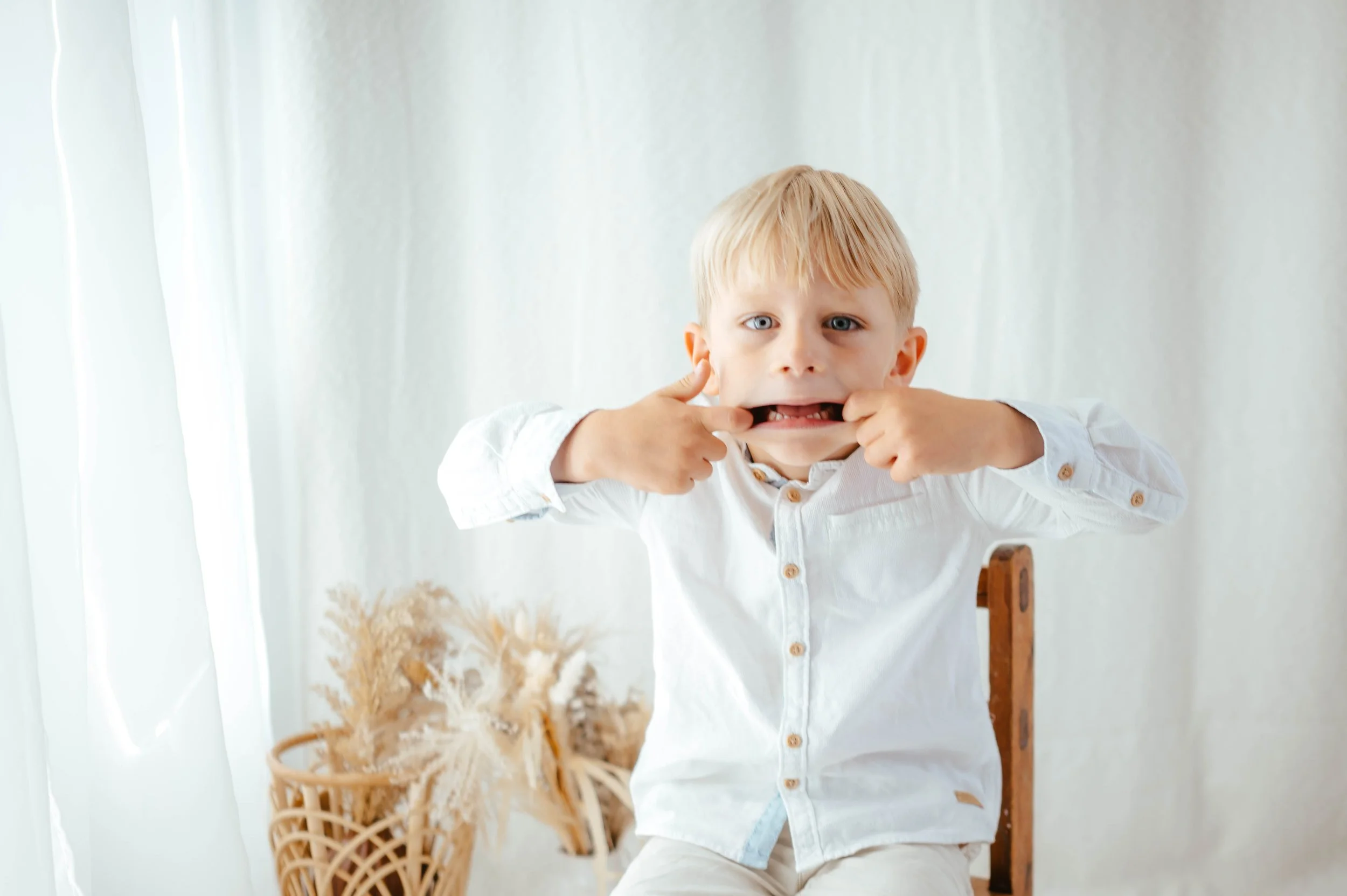 A young boy with blonde hair and blue eyes making a funny face by pulling his lips with his fingers, sitting in front of a white curtain with dried plants in a basket nearby.