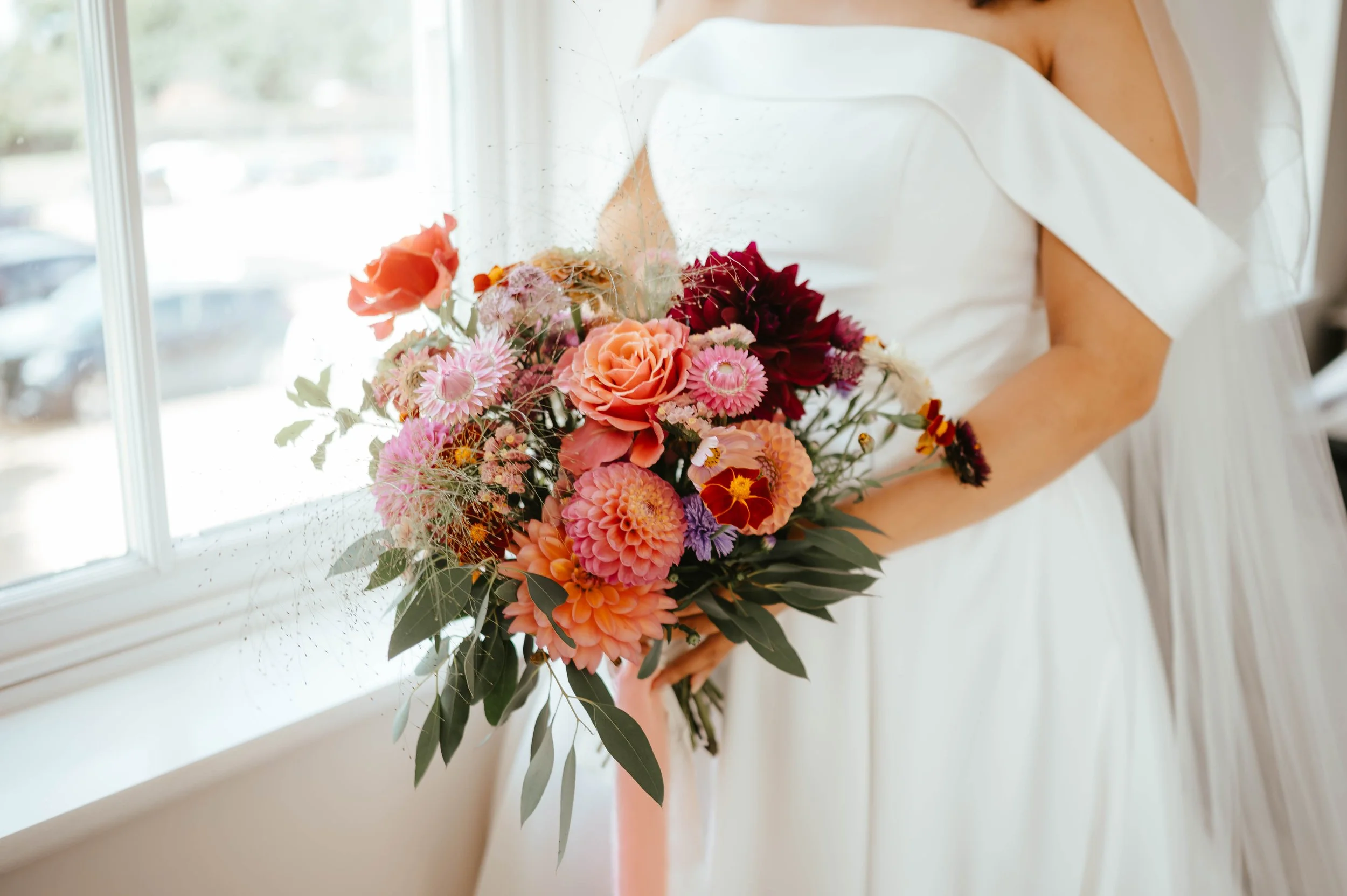 A bride holding a bouquet of pink, orange, and red flowers near a window.