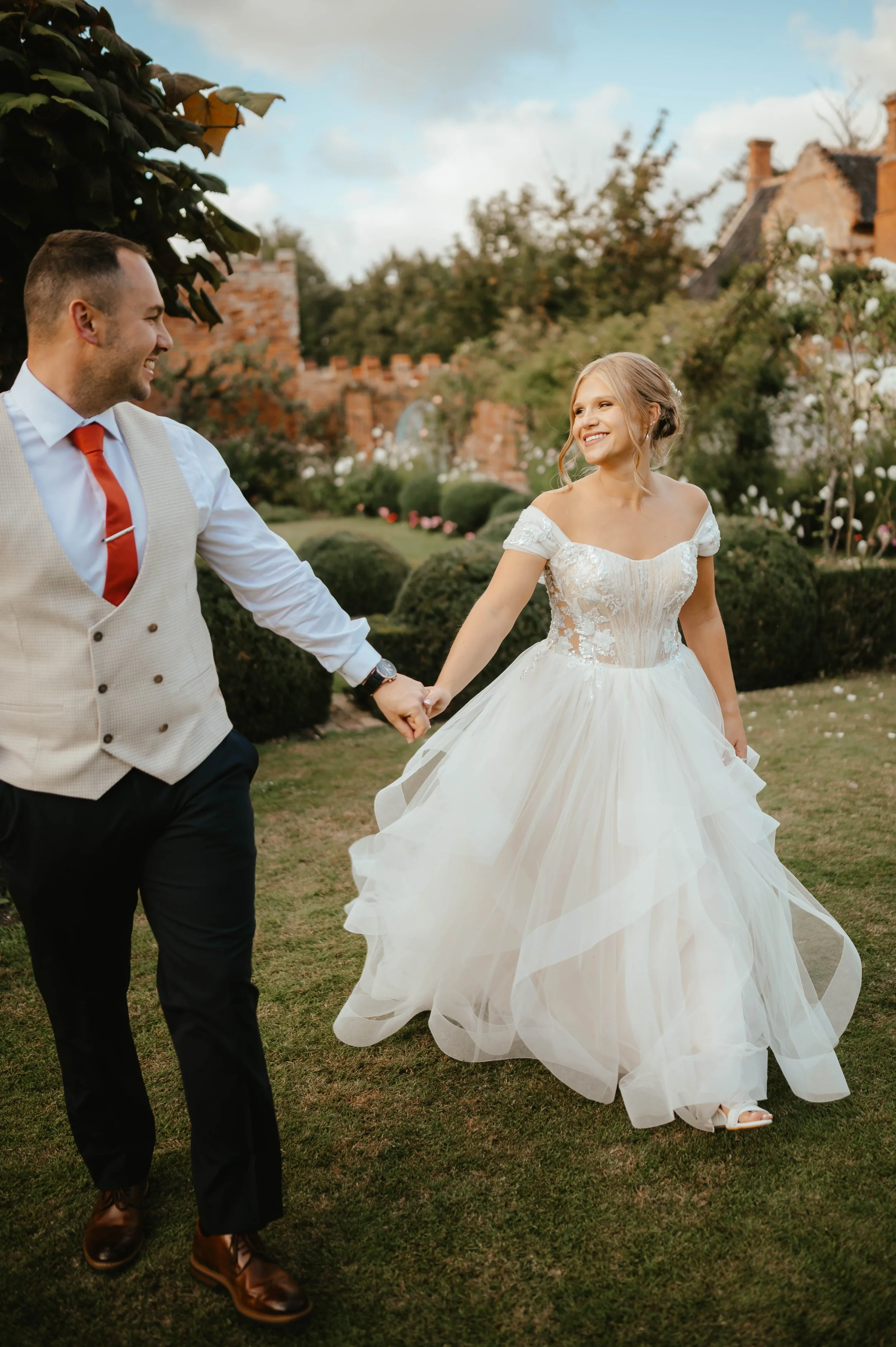 A newlywed couple holding hands and smiling in a garden with a brick building in the background.