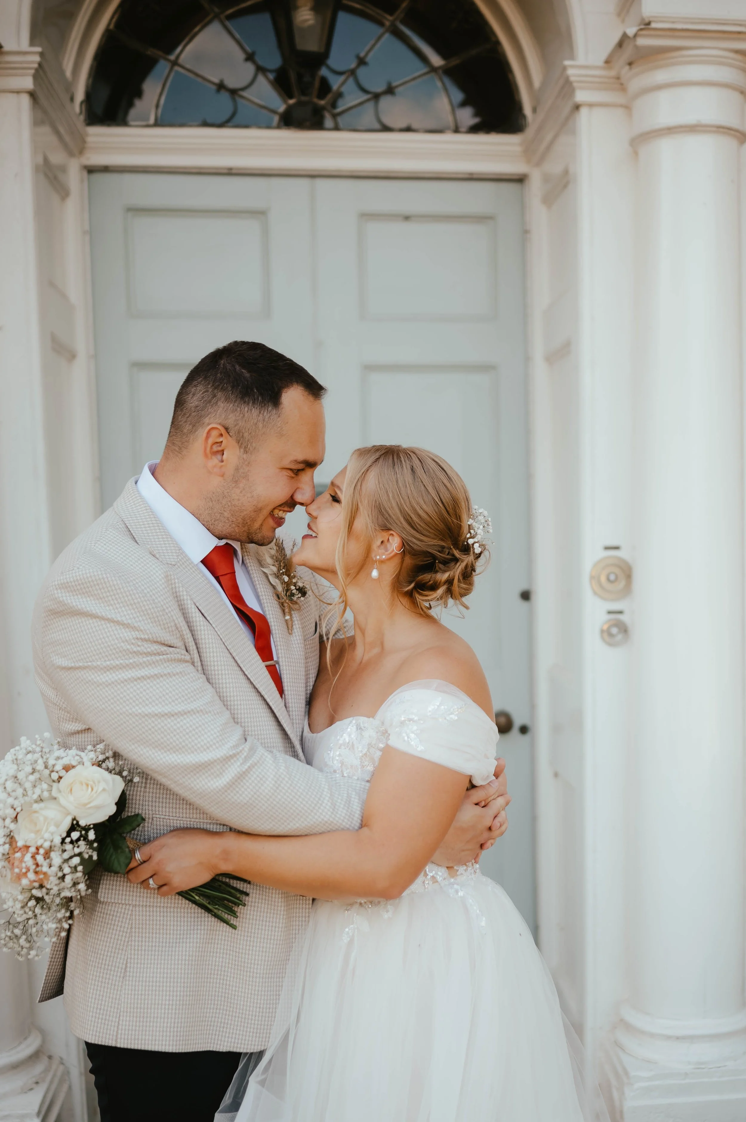 A bride and groom embrace in front of a white door, smiling and touching noses affectionately.