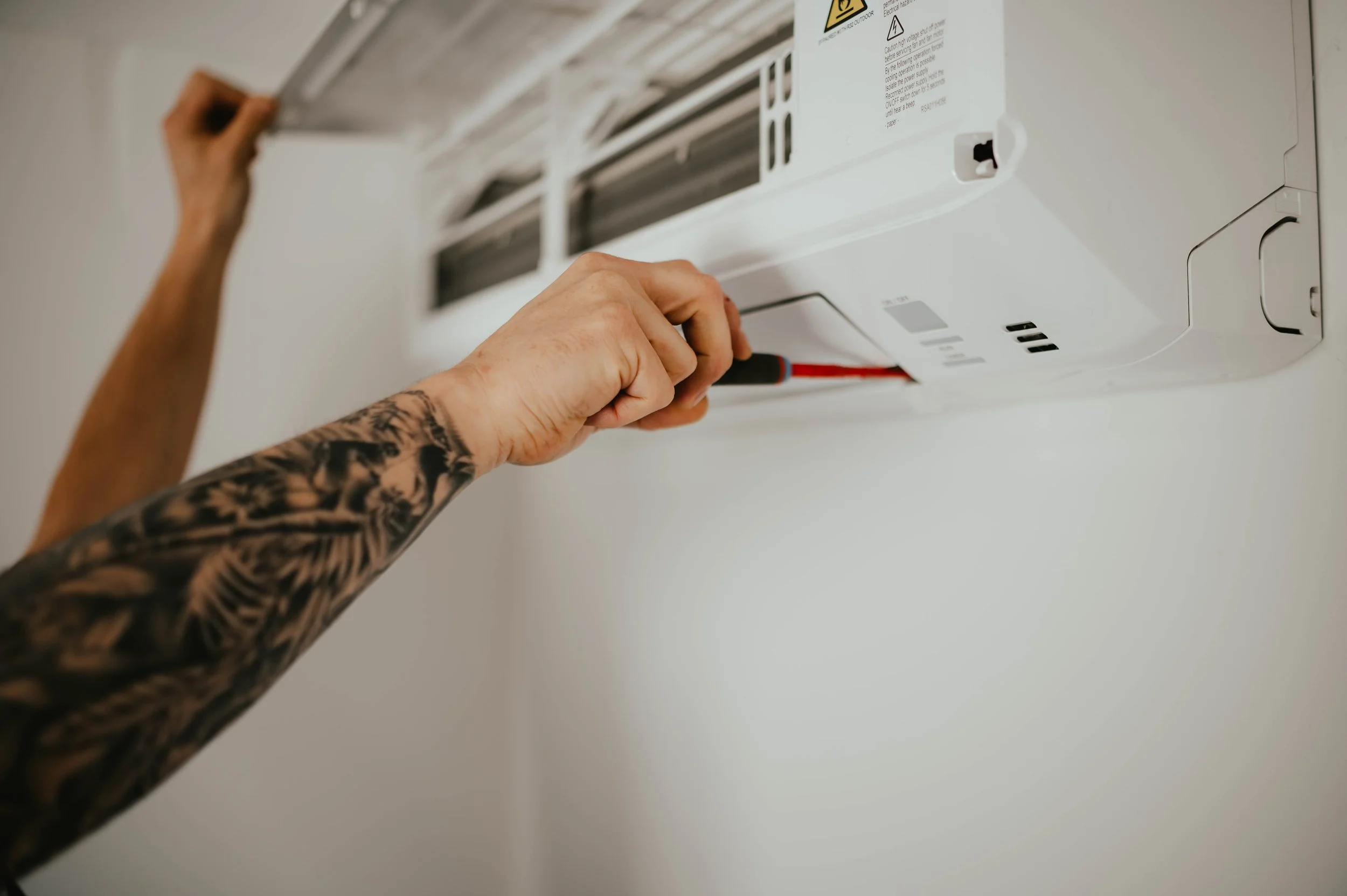 A person with a tattooed arm fixing or servicing a white wall-mounted air conditioning unit using a screwdriver.