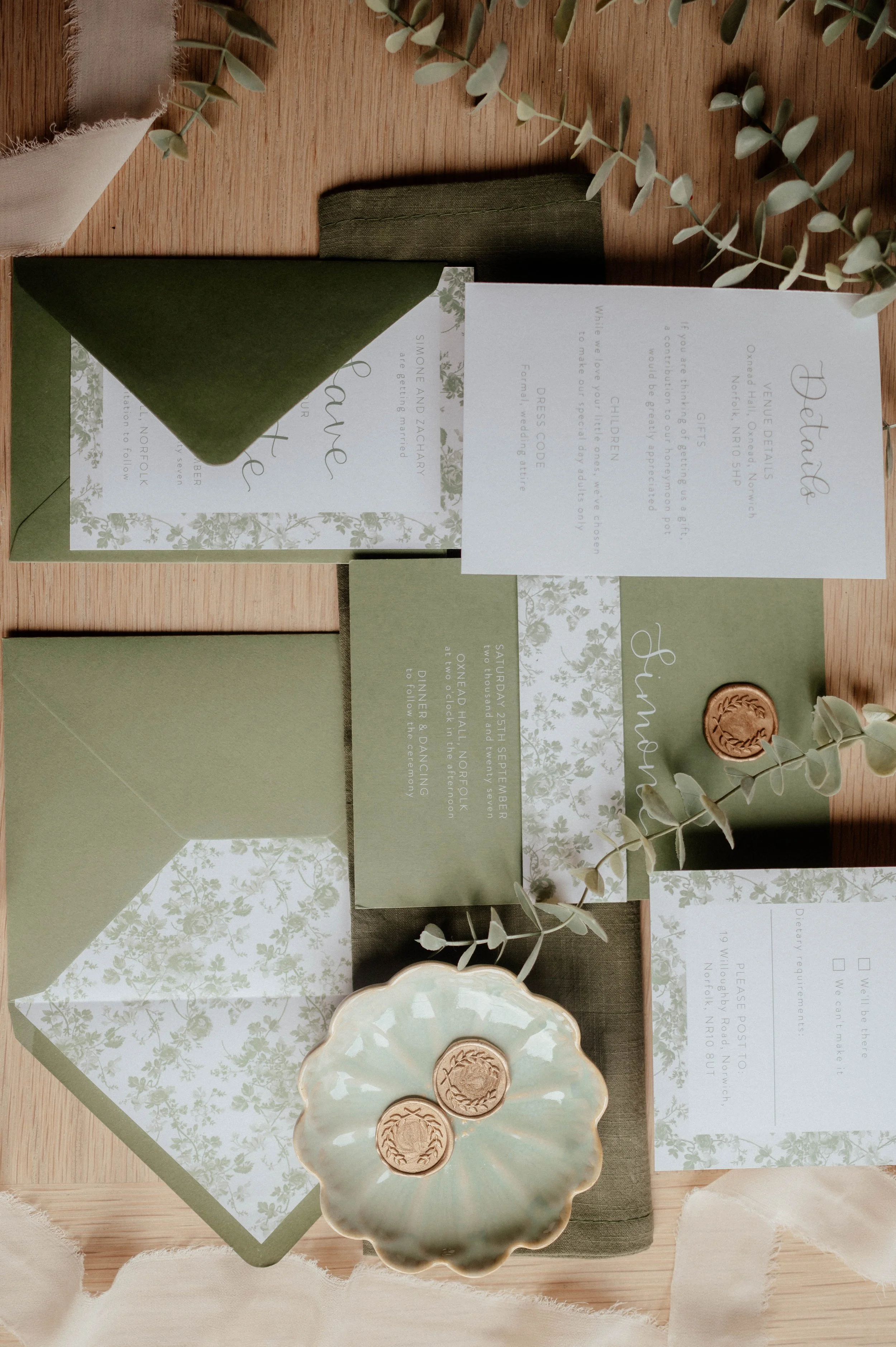 Invitation suite with green and white floral accents, envelope, and sealing wax on a wooden table, decorated with eucalyptus leaves.