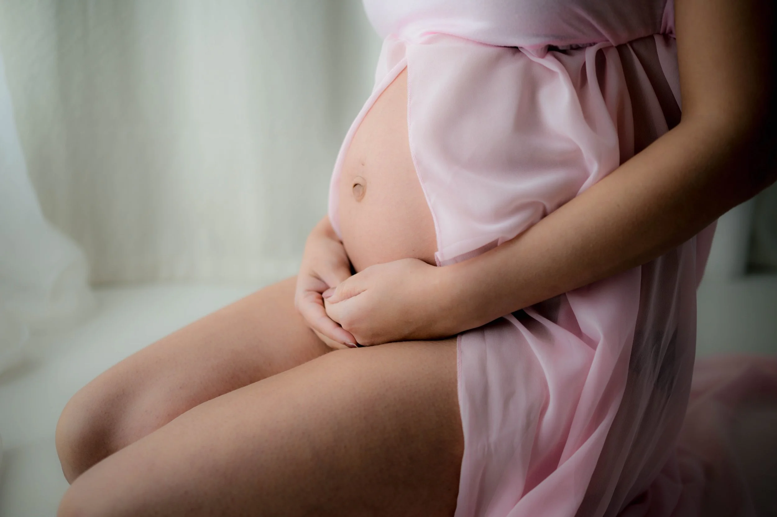 Pregnant woman in pink silk nightgown sitting on bed, holding her belly.