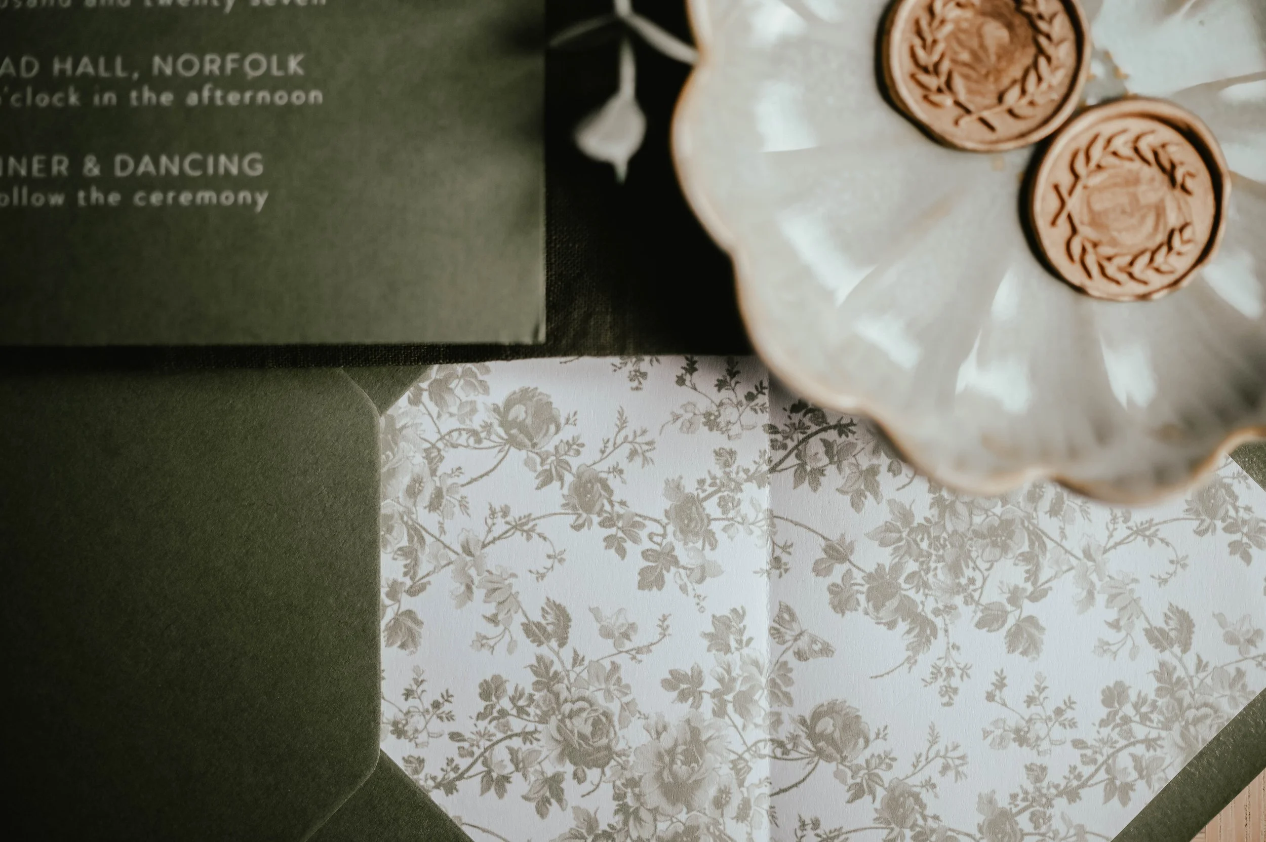 Partially visible green event program or invitation with white text, a white floral-patterned napkin or tablecloth, and a white decorative cake stand holding two small chocolate coins in paper wrappers with a crest design.