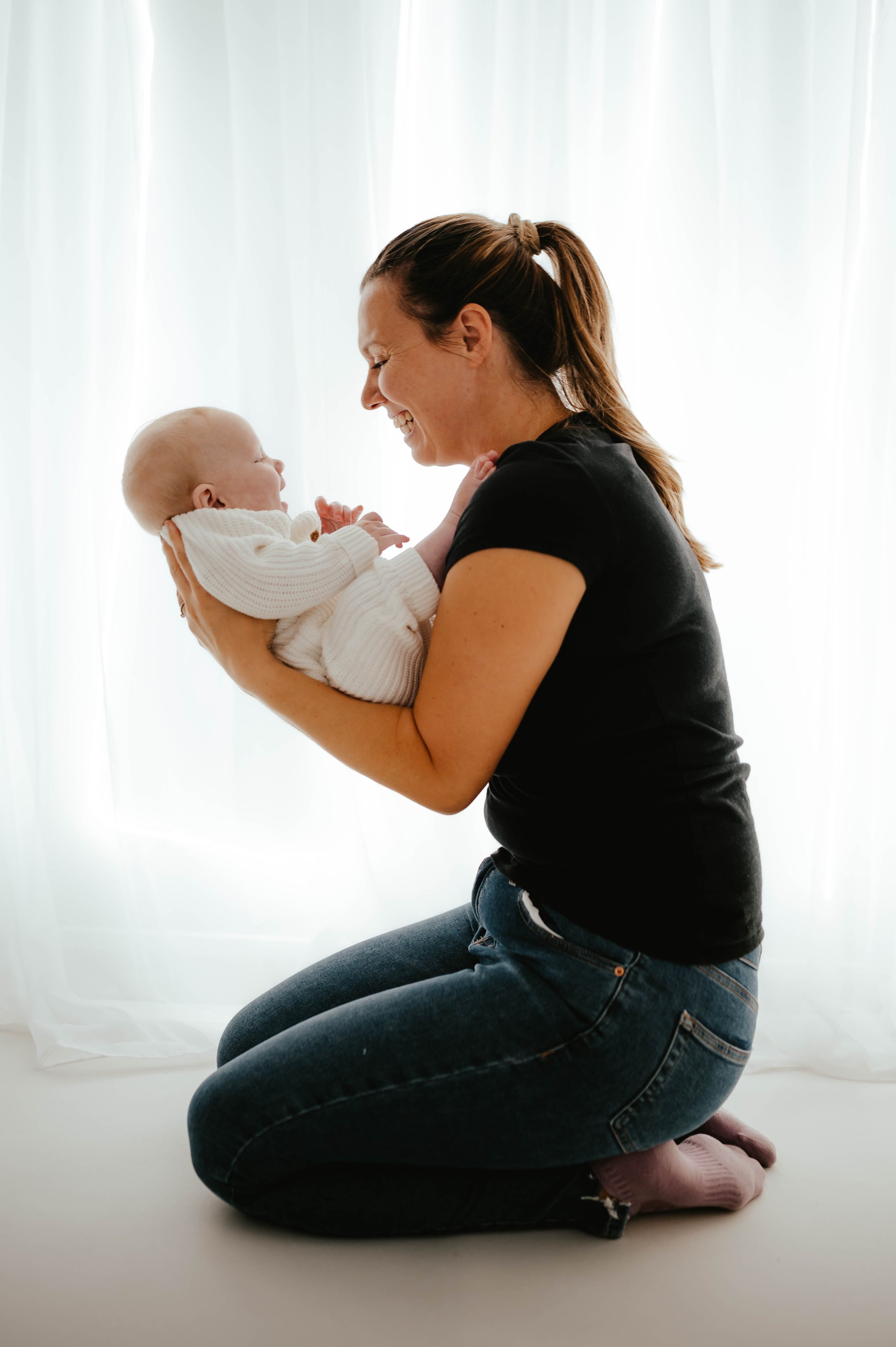 A woman kneeling and holding a baby close to her face, both smiling at each other, in front of a sheer curtain background.
