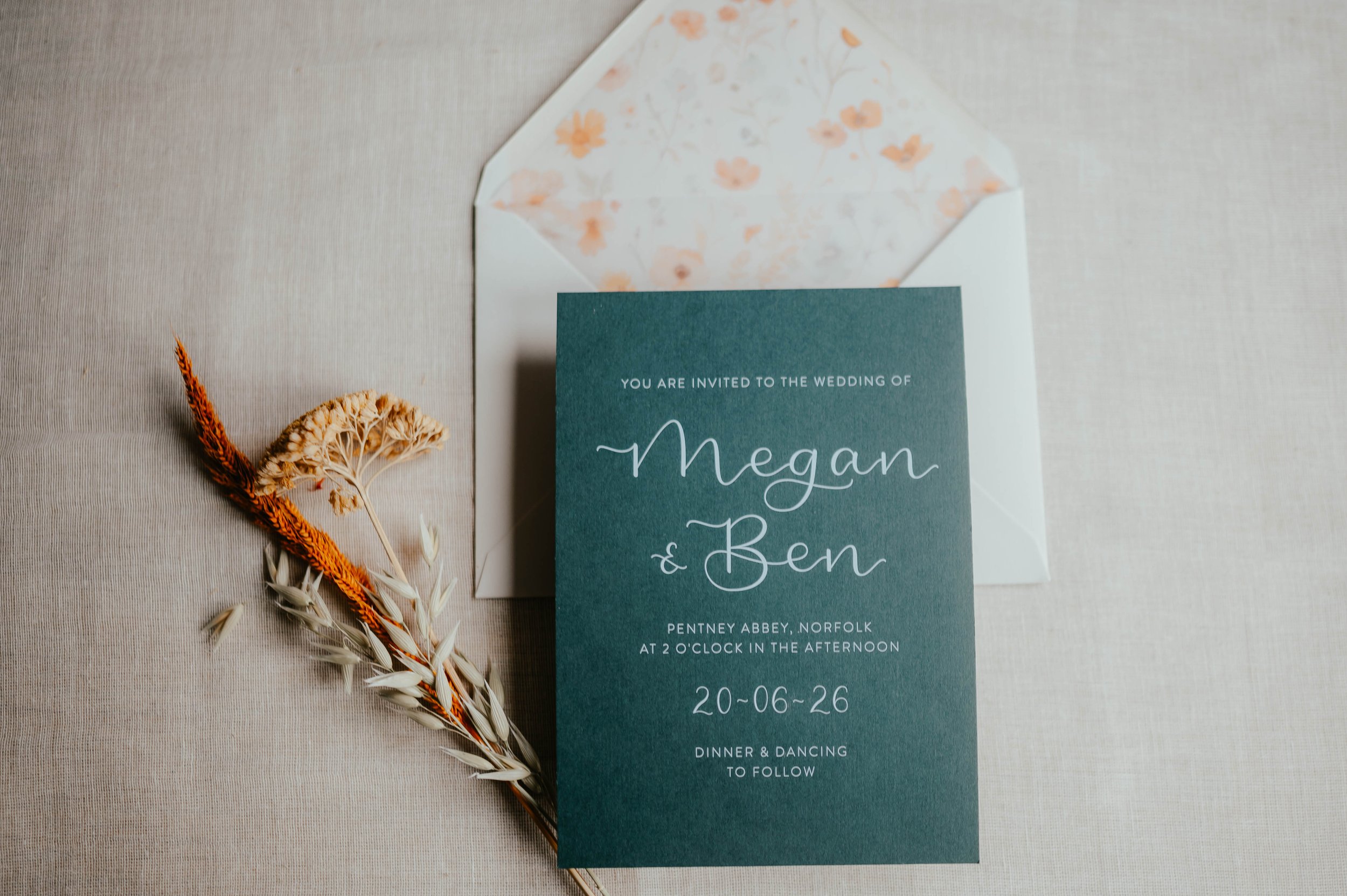 Wedding invitation card for Megan and Ben placed on a light-colored fabric surface. The card is teal with white text, and there is an open white envelope with a floral pattern lining behind it. Dried flowers are positioned to the left of the card.