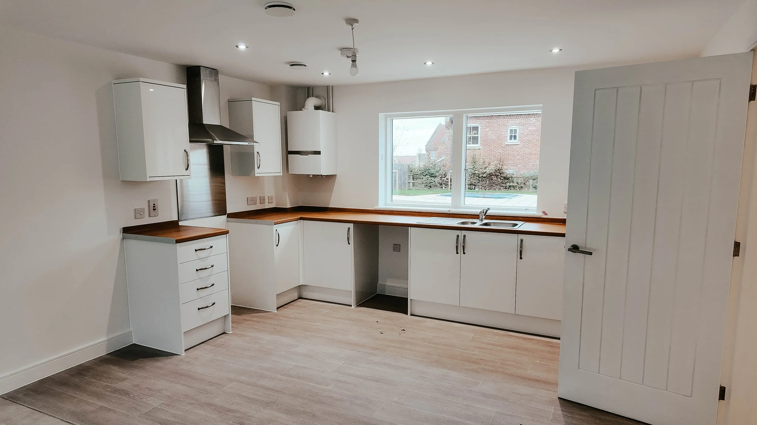 Empty kitchen with white cabinets, wooden countertops, a window overlooking a backyard, and a door open to the right.
