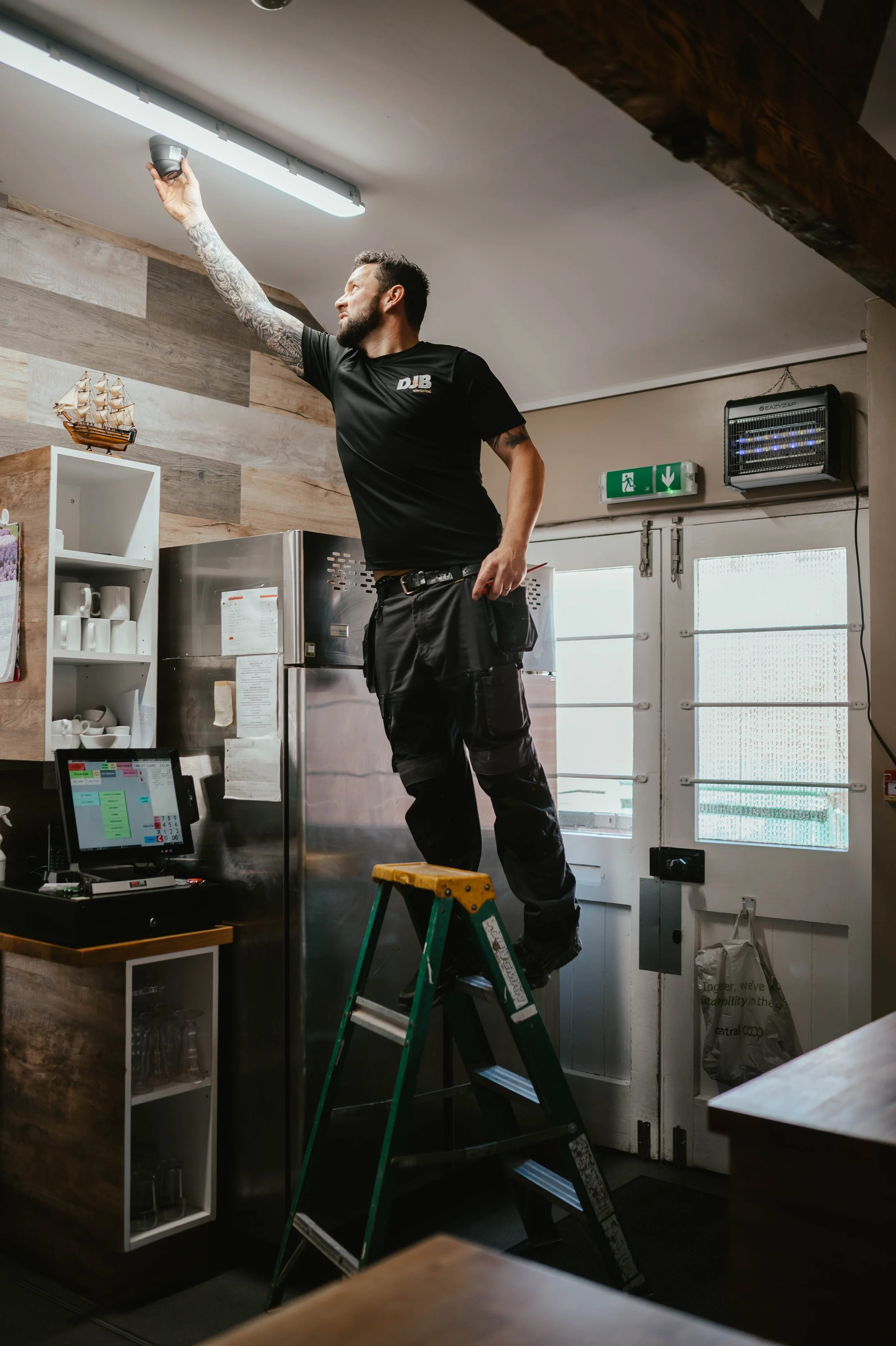 A man with tattoos on his arm standing on a ladder cleaning a ceiling light fixture in a restaurant or cafe.