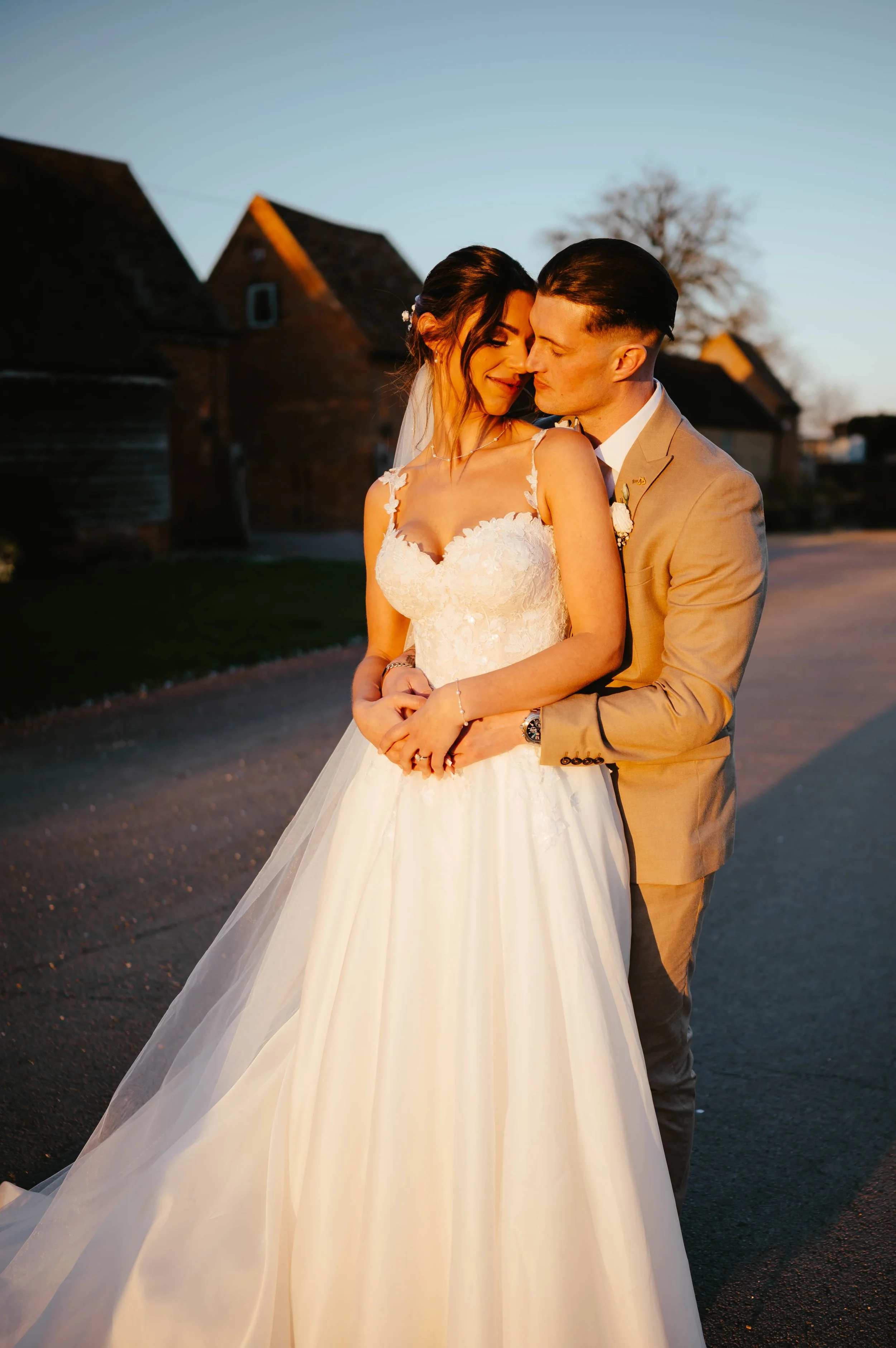 A newlywed couple embracing outdoors during sunset, with rustic buildings in the background.