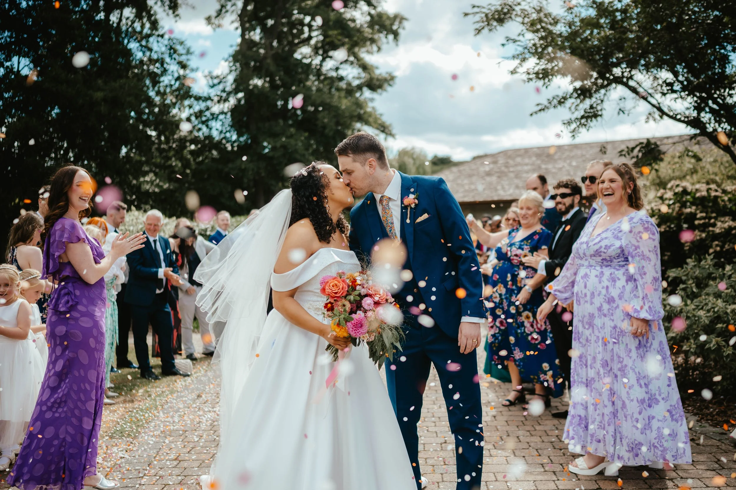 A newlywed couple sharing a kiss during their wedding ceremony, surrounded by family and friends throwing colorful confetti outdoors.