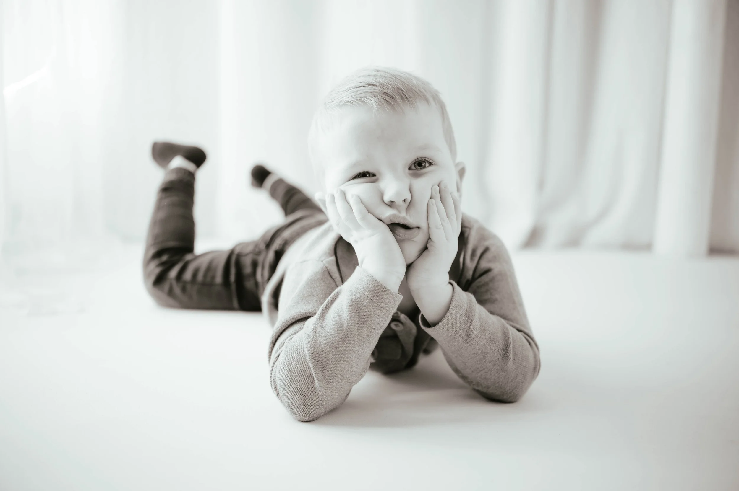A young boy with short hair lying on his stomach on a bed, resting his face in his hands, looking at the camera with a curious expression.