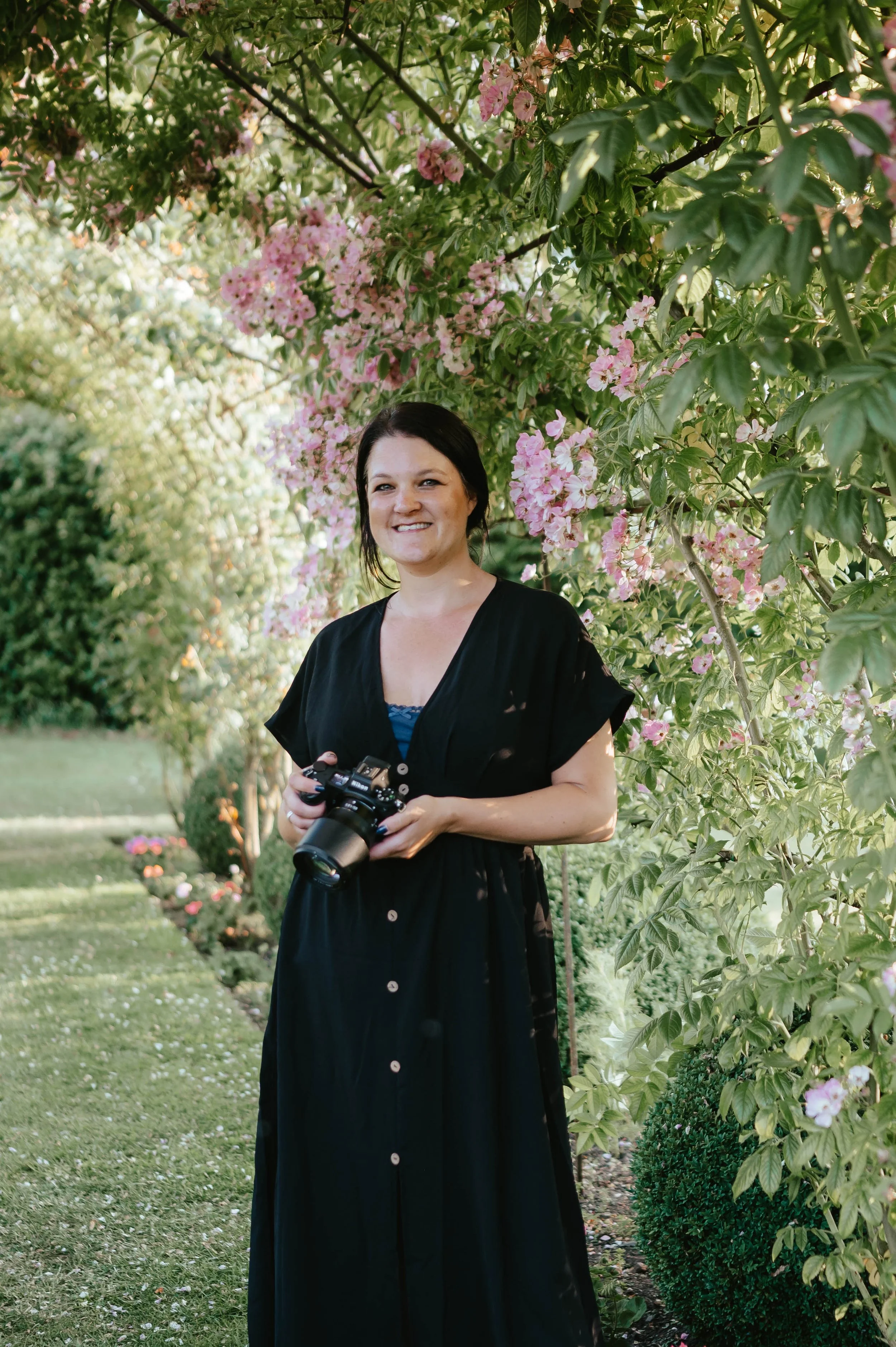 A woman with dark hair wearing a black dress and holding a camera, standing outdoors in a garden with pink flowers and green foliage.