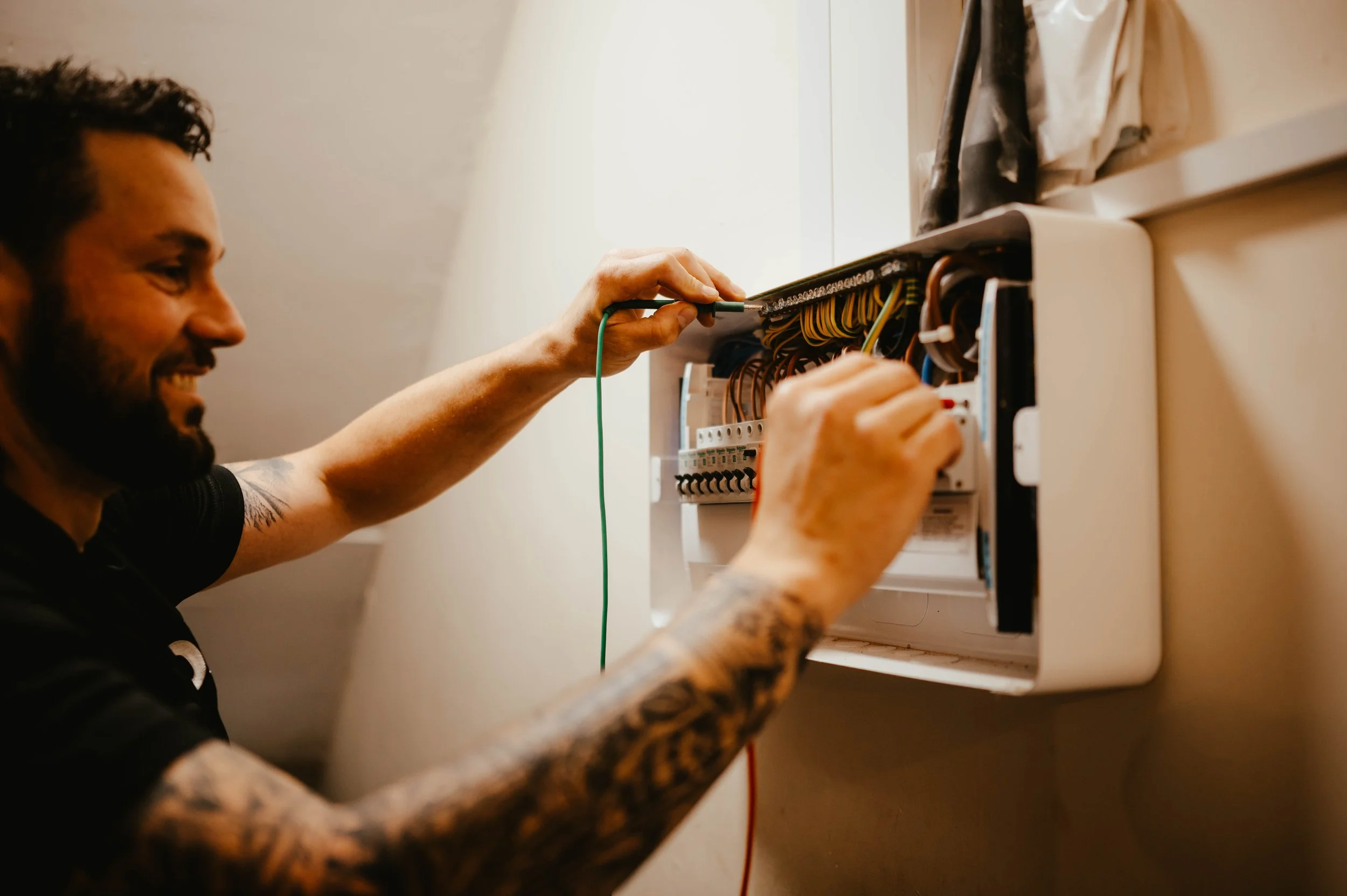 Man working on electrical panel, reaching inside with tools, smiling, tattoos on arms, beige wall background.