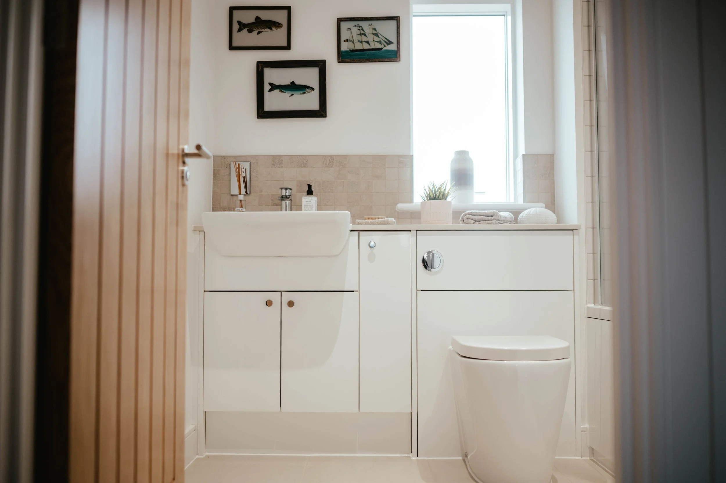 A small, modern bathroom with a white vanity, a sink, and a white toilet. The vanity has a cabinet with two doors and a small drawer. On top of the vanity are a soap dispenser, a toothbrush holder, and a small tray. Above the vanity are framed photos
