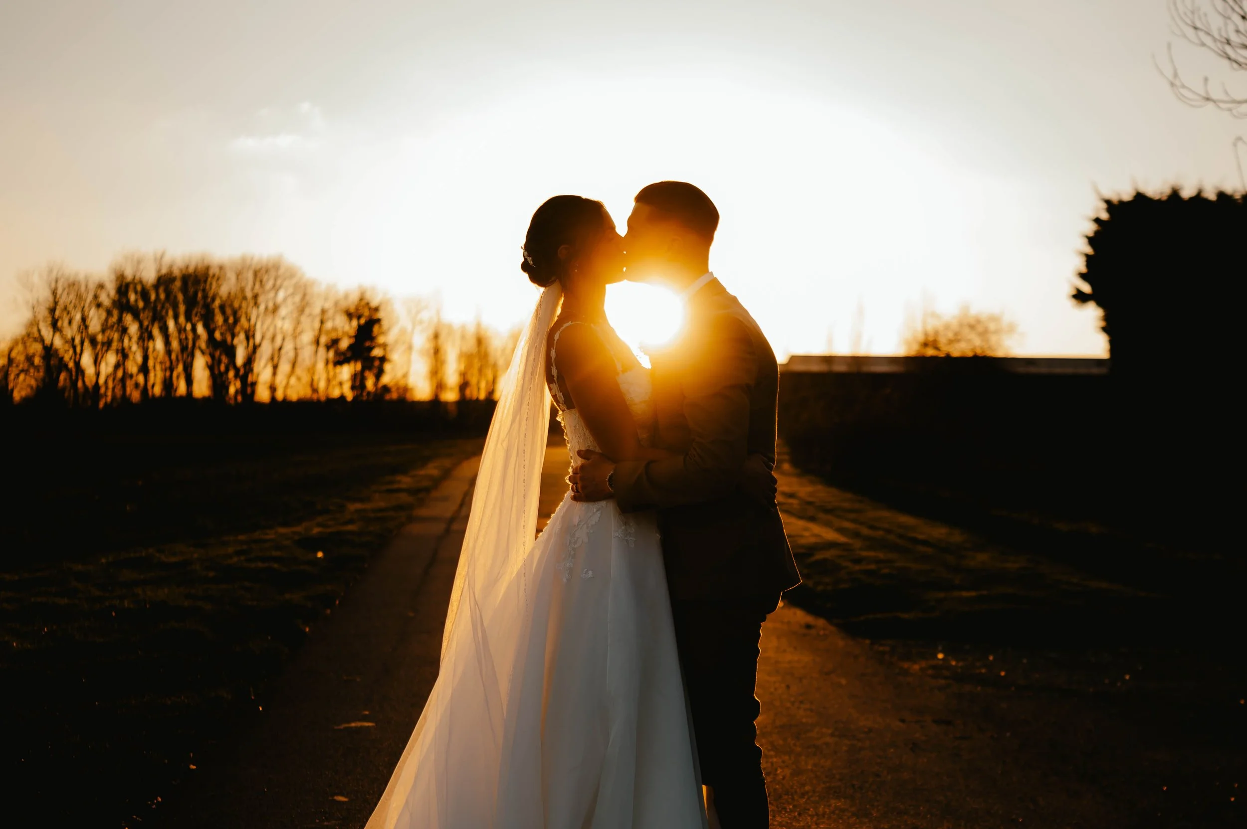 Silhouette of a newlywed couple kissing at sunset, standing on a path with trees in the background.