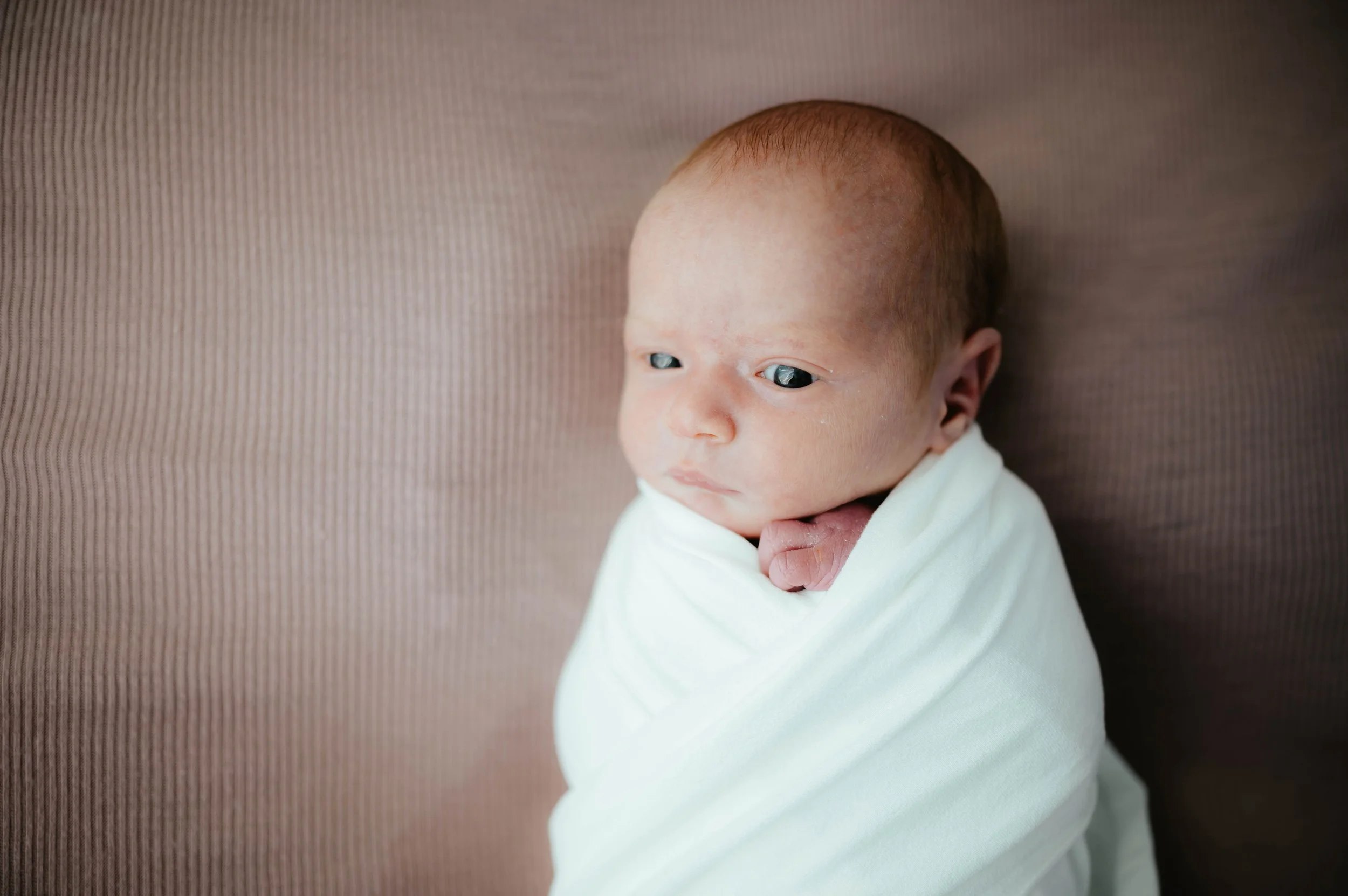 Newborn baby wrapped in a white blanket, lying on a brown textured surface, looking to the side.