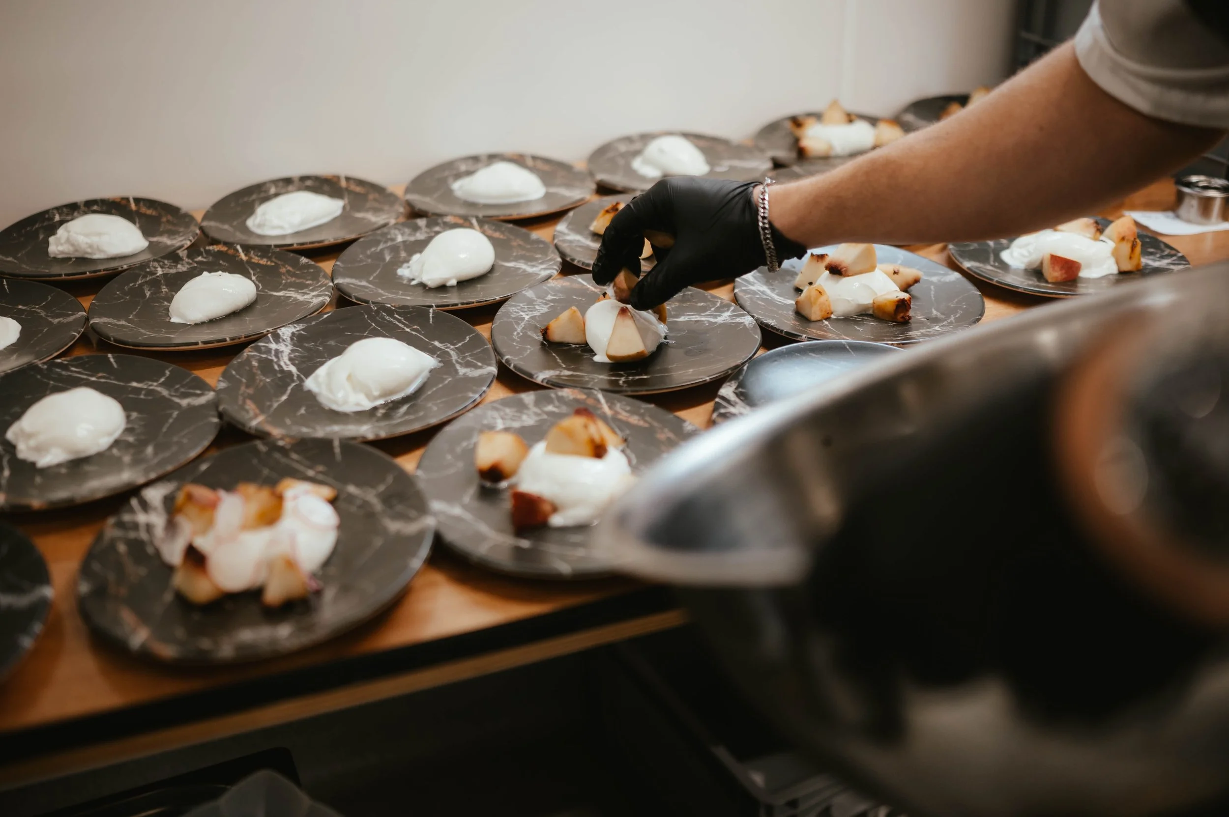 A person wearing a white shirt and black glove arranging small dessert portions on black marble-patterned plates, with some plates topped with a white dollop and others with cut fruit, on a wooden table.