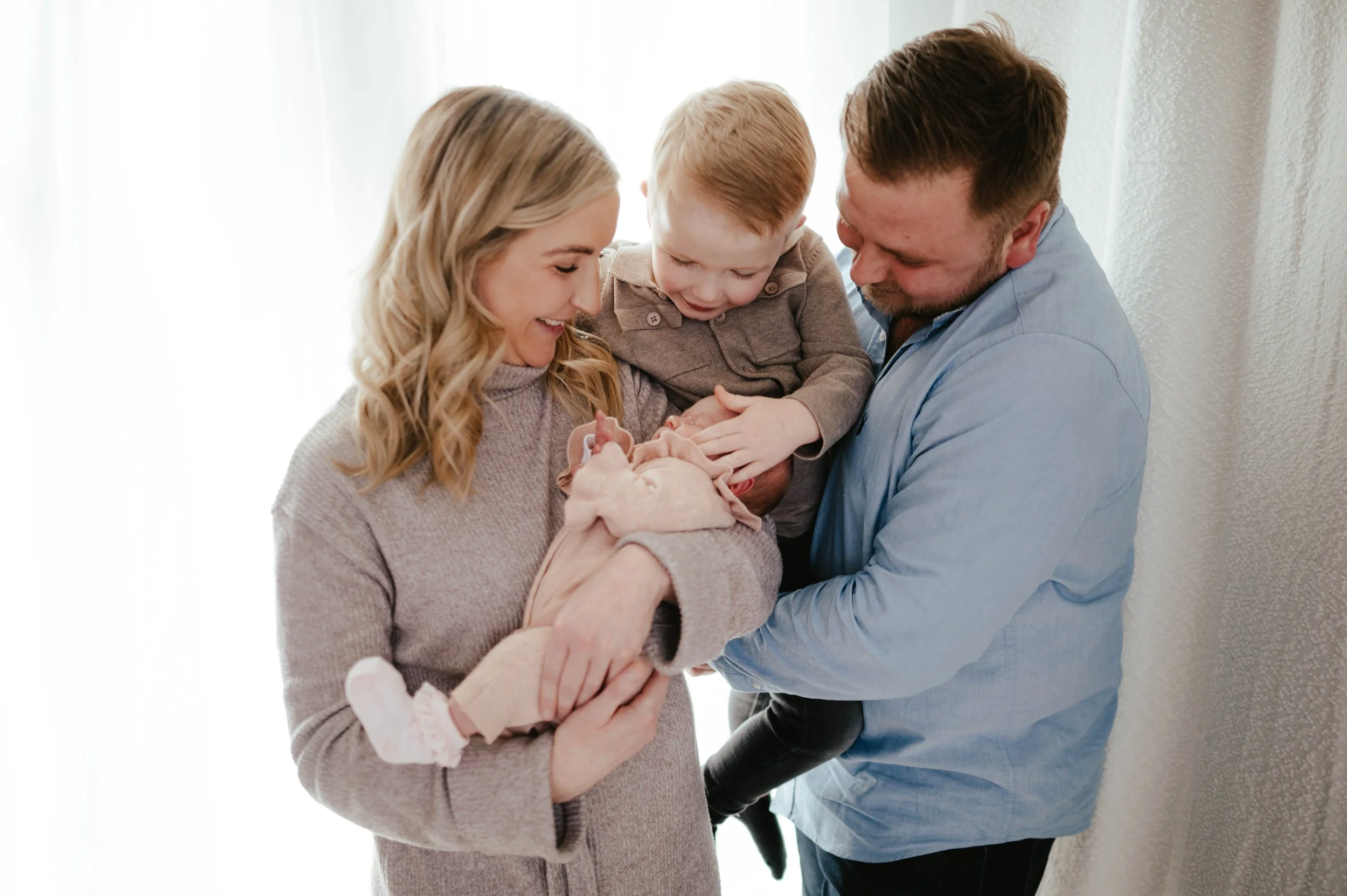 A family of four, including a mother, father, young boy, and newborn baby, gathered around and smiling as they look at the newborn, in a bright room with natural light.