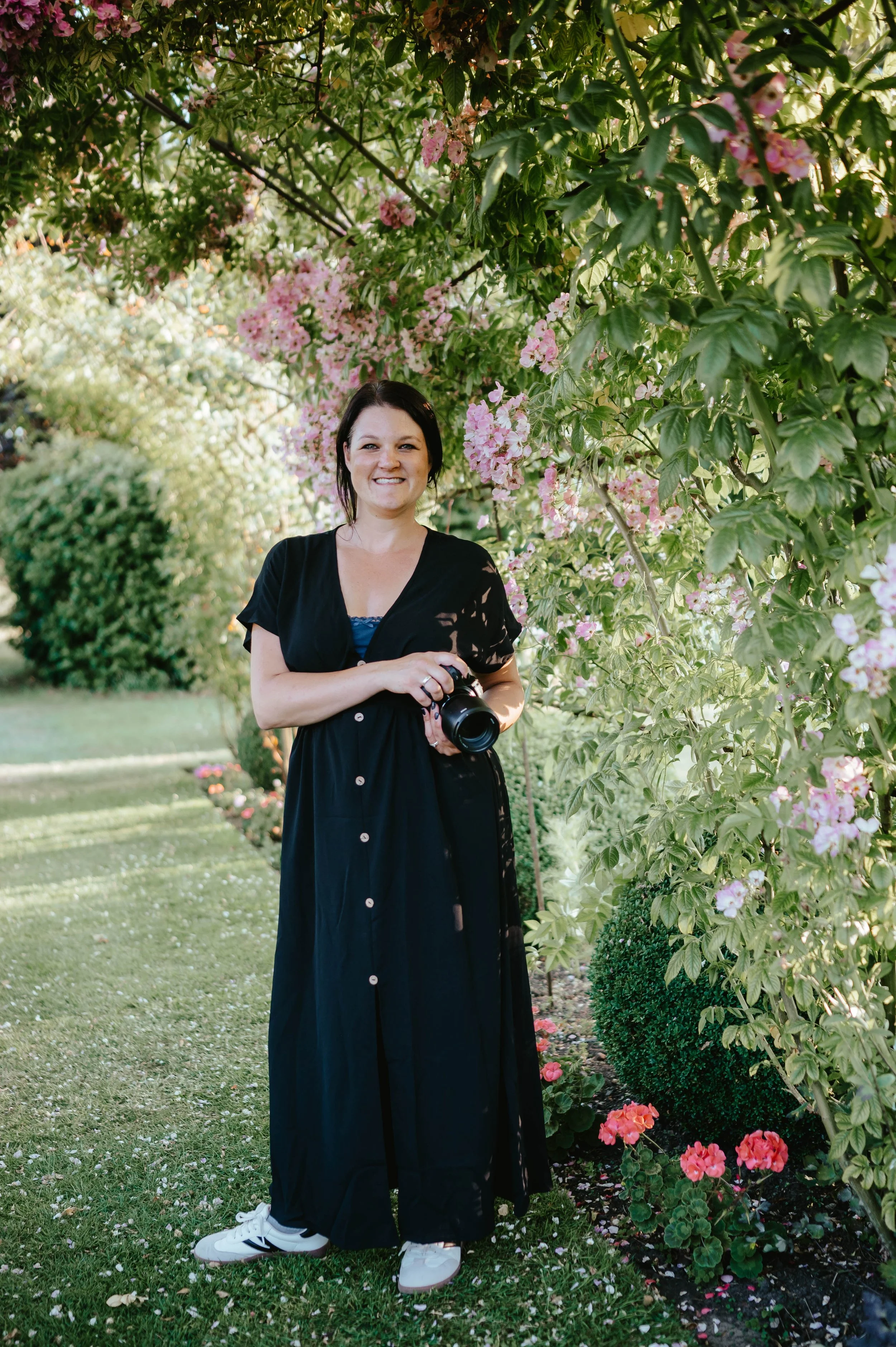 A woman standing in a garden with pink and white flowers and greenery, holding a camera, smiling.