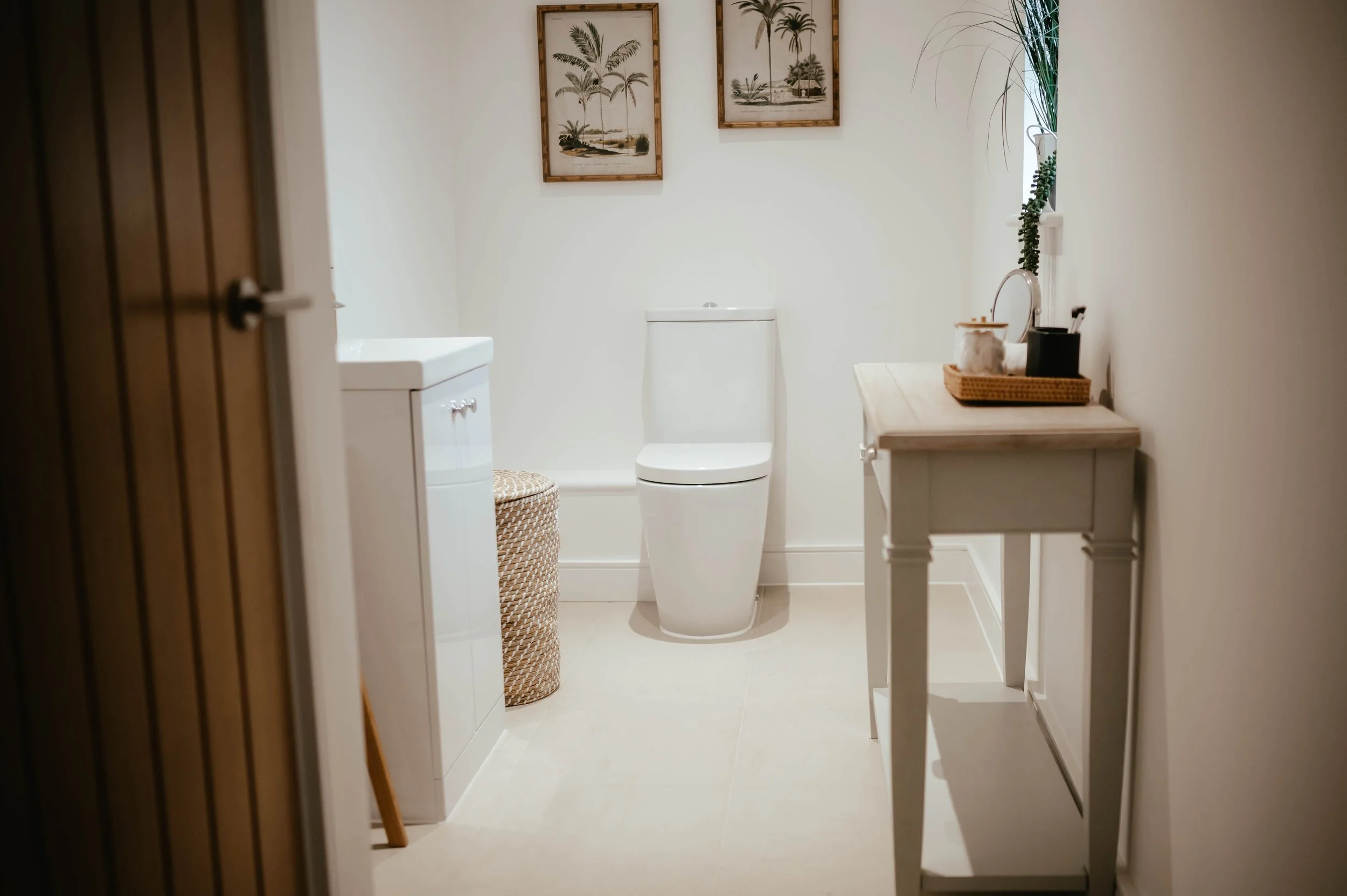 A clean and minimal bathroom with a white toilet, a small white cabinet, a wicker laundry basket, framed beach-themed artwork on the wall, and a wooden table with decorative items near a window with green plants.