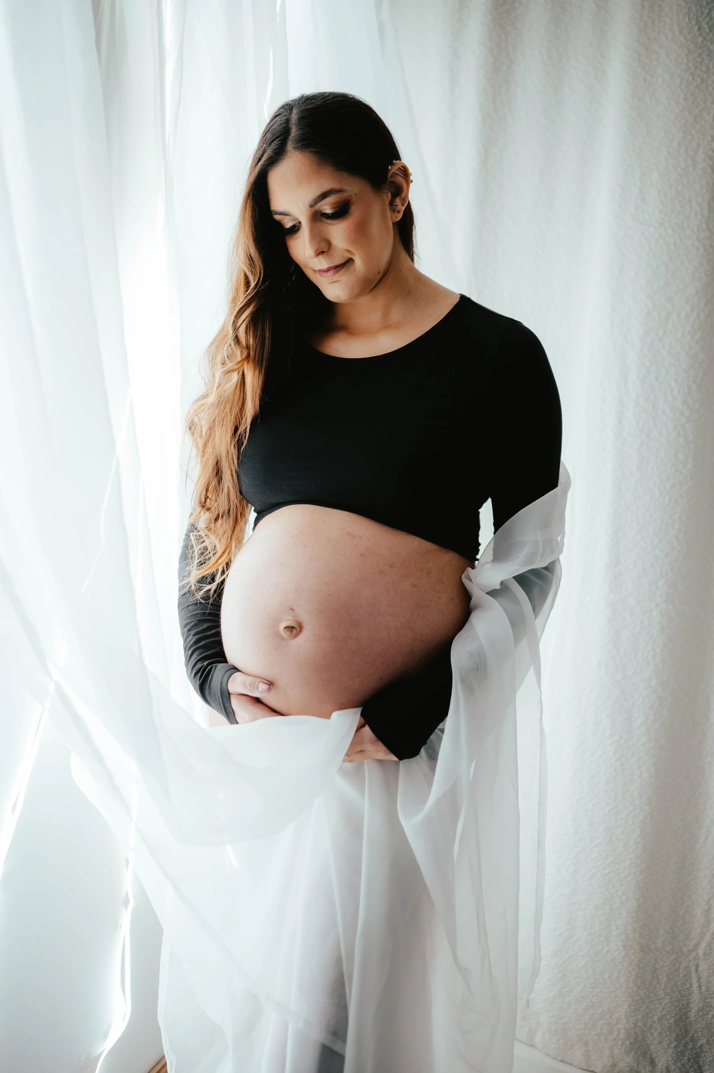 Pregnant woman in black top with white fabric over her arm, standing by a window with white curtains, looking down at her belly.