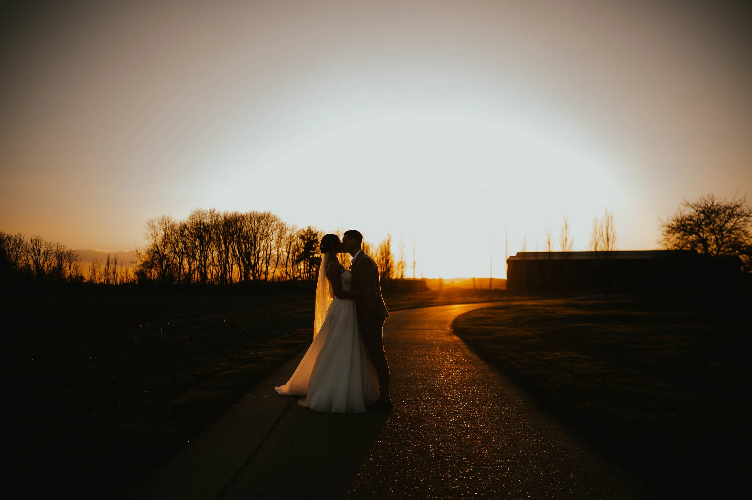 A silhouette of a newlywed couple holding hands and kissing on a path during sunset, with trees and a building in the background.