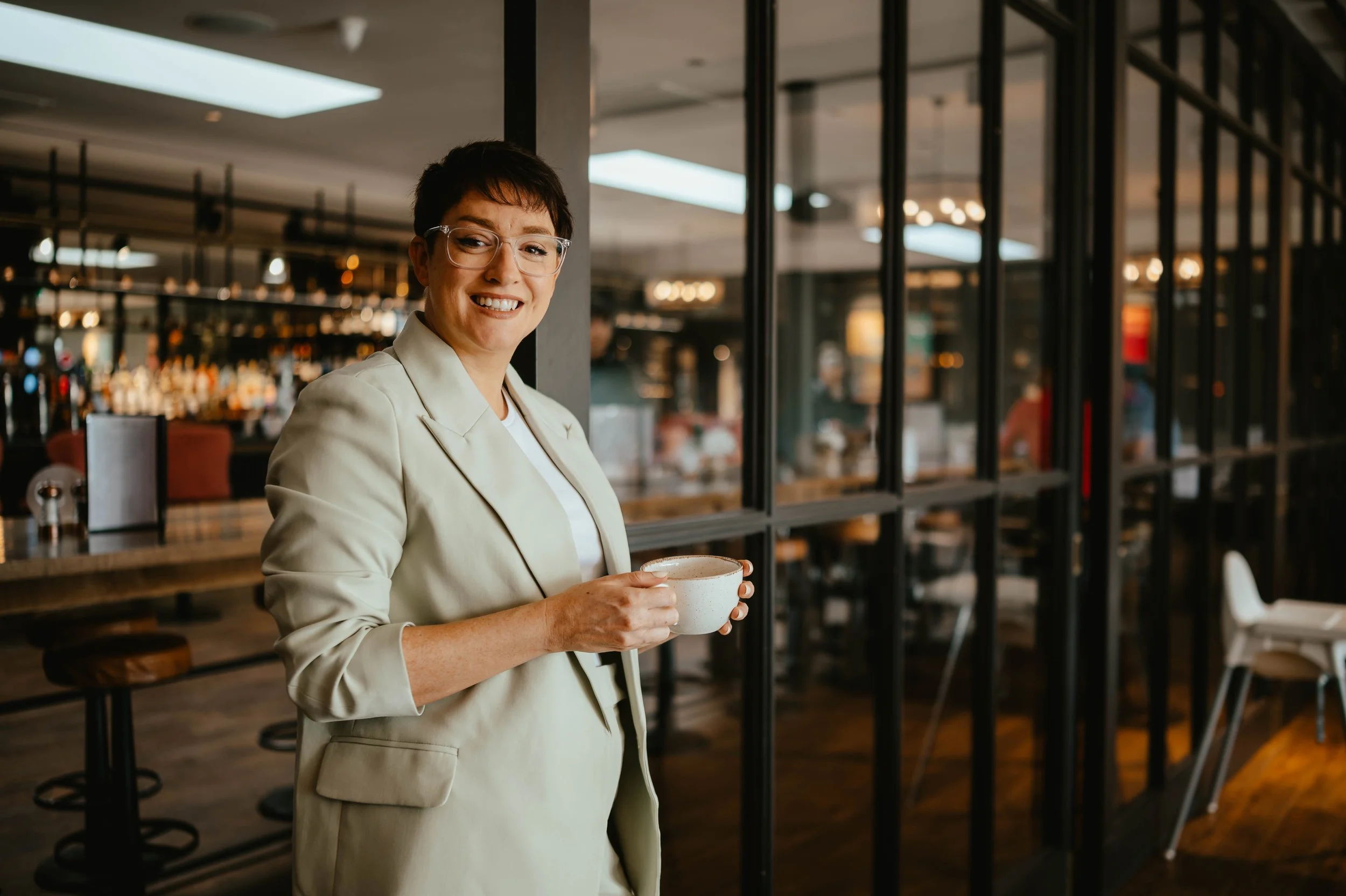 A woman with short dark hair and glasses smiling and holding a coffee cup inside a modern cafe with wood and black metal decor.