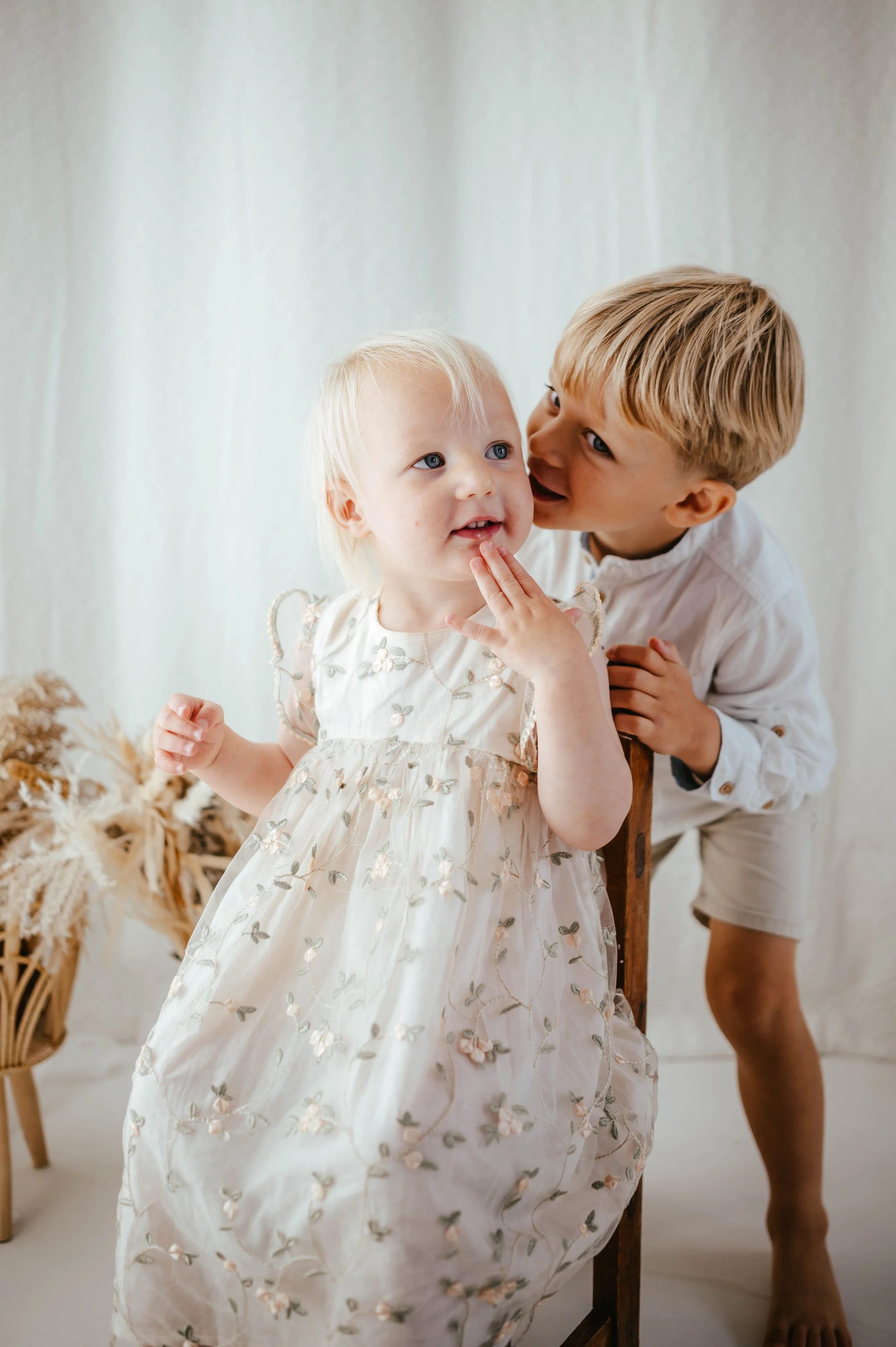 A young boy whispering to a young girl with blonde hair, wearing a white embroidered dress, in a bright indoor setting with dried flowers in the background.