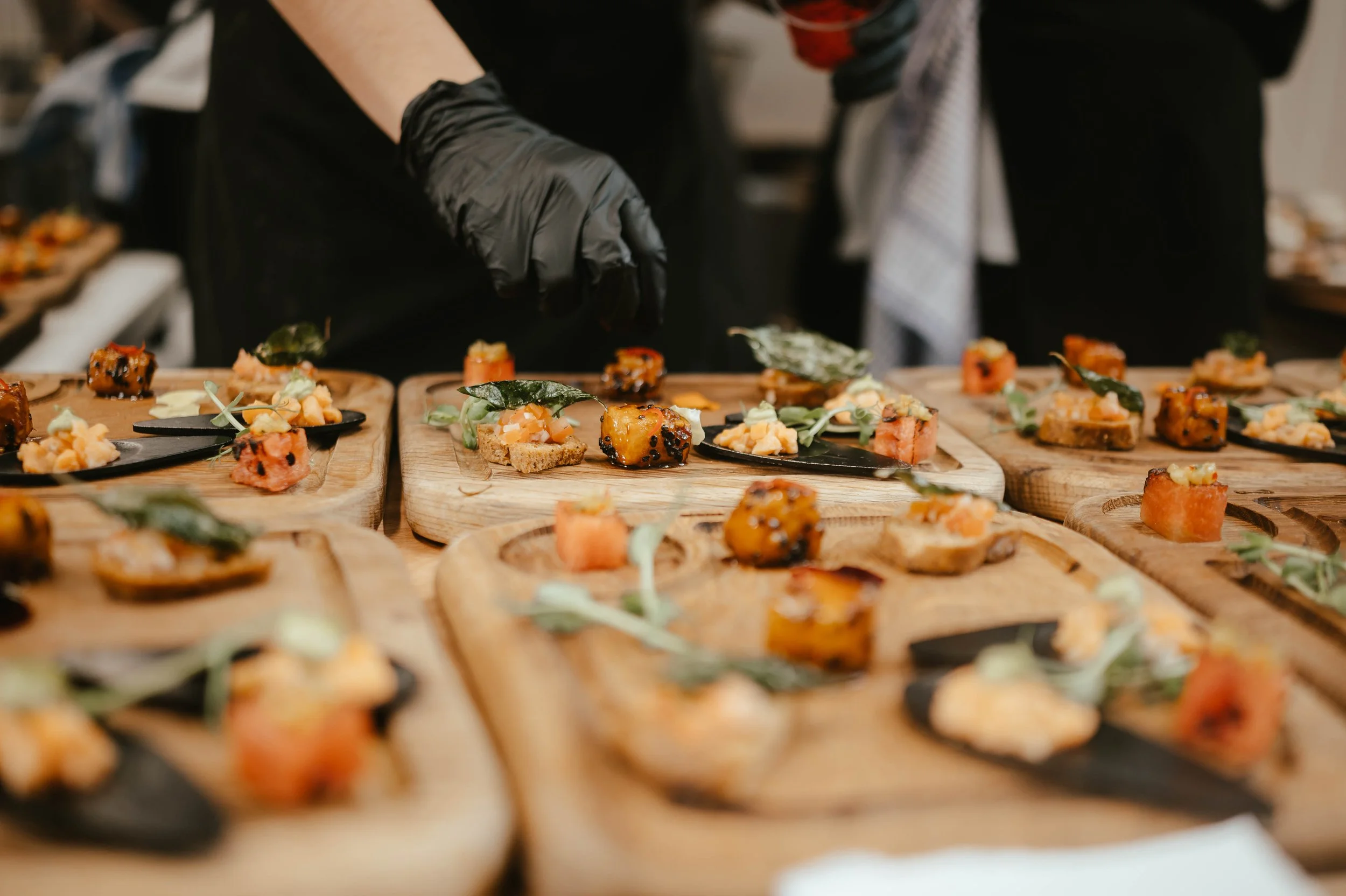A person wearing black gloves preparing assorted canapés on wooden serving boards, with various toppings and garnishes.