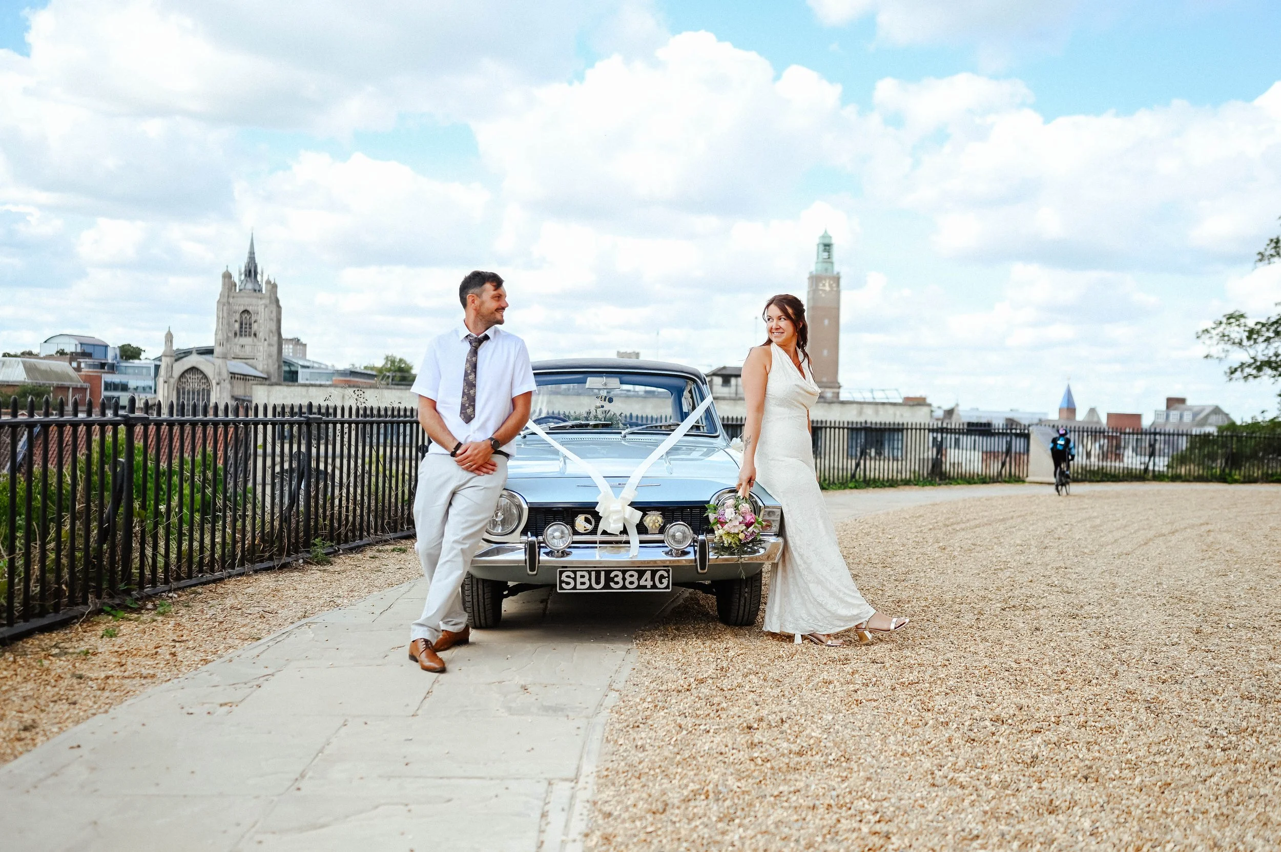 A bride and groom posing near a decorated vintage car outdoors on a cloudy day, with city buildings and a fence in the background.