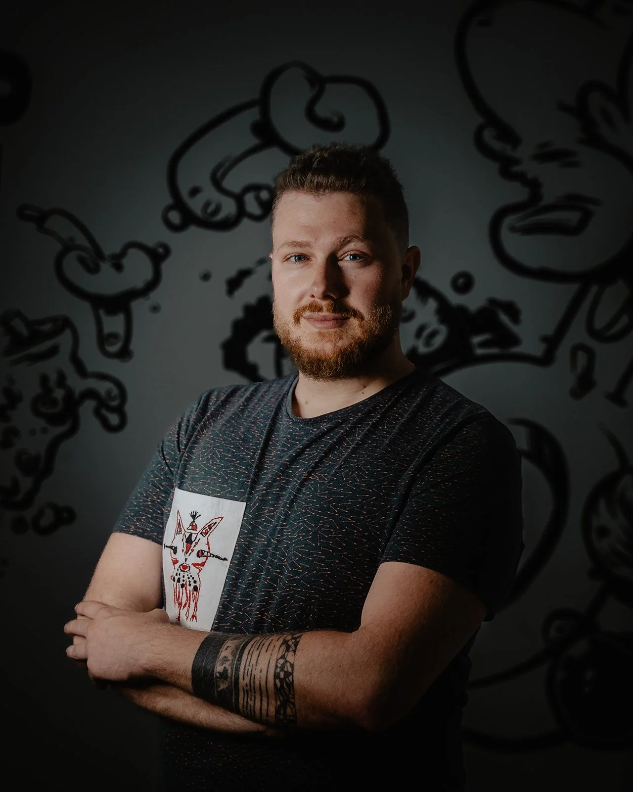 A young man with red hair and a beard crossing his arms, wearing a black shirt with a graphic of a goat in the center. The background features abstract black line art on a dark wall.