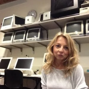 A woman with blonde hair in a white top sitting in an office or tech room with old computers and monitors on shelves behind her.