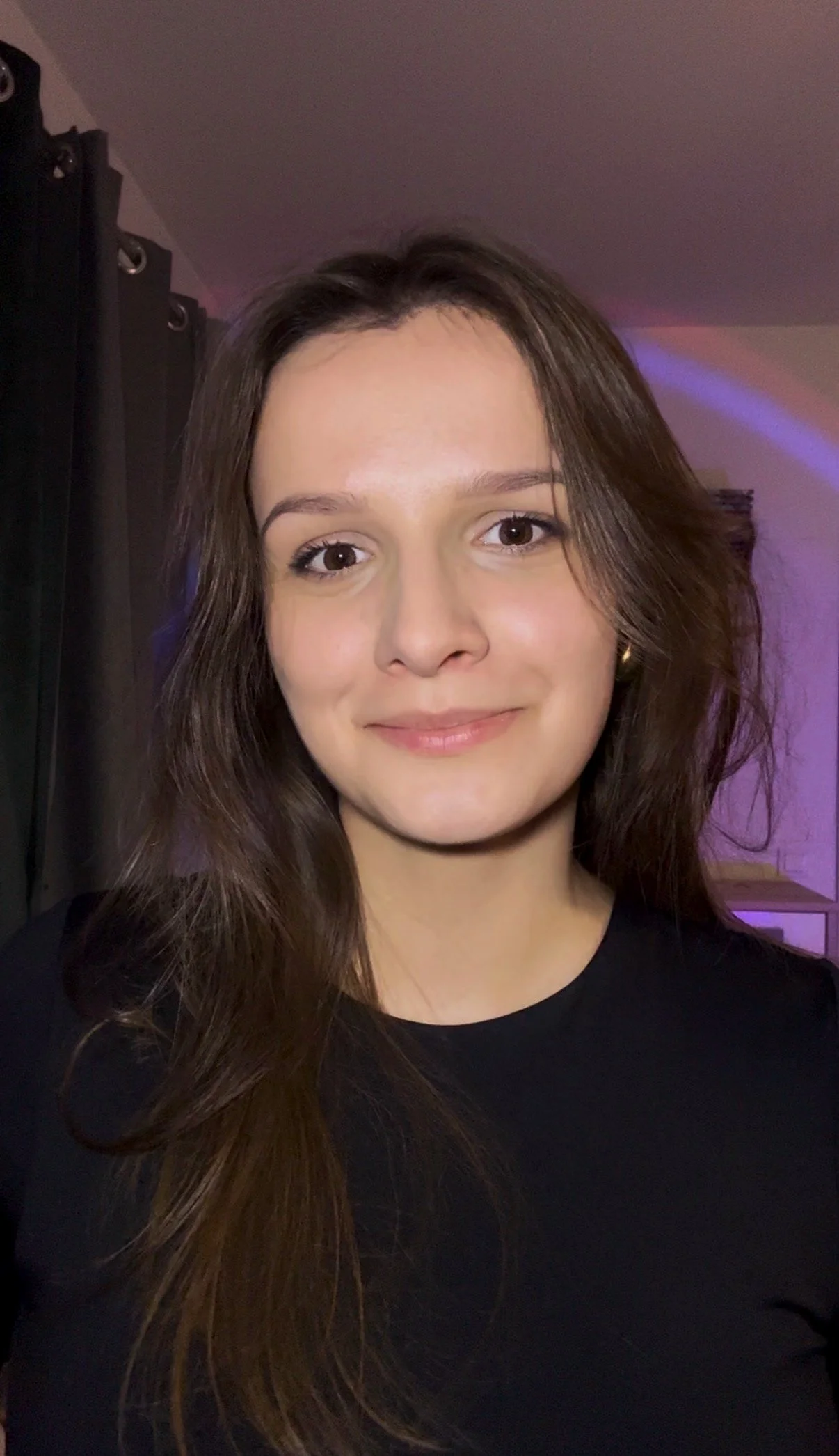 A young woman with brown hair wearing a black top, smiling in a room with dark curtains and purple lighting.