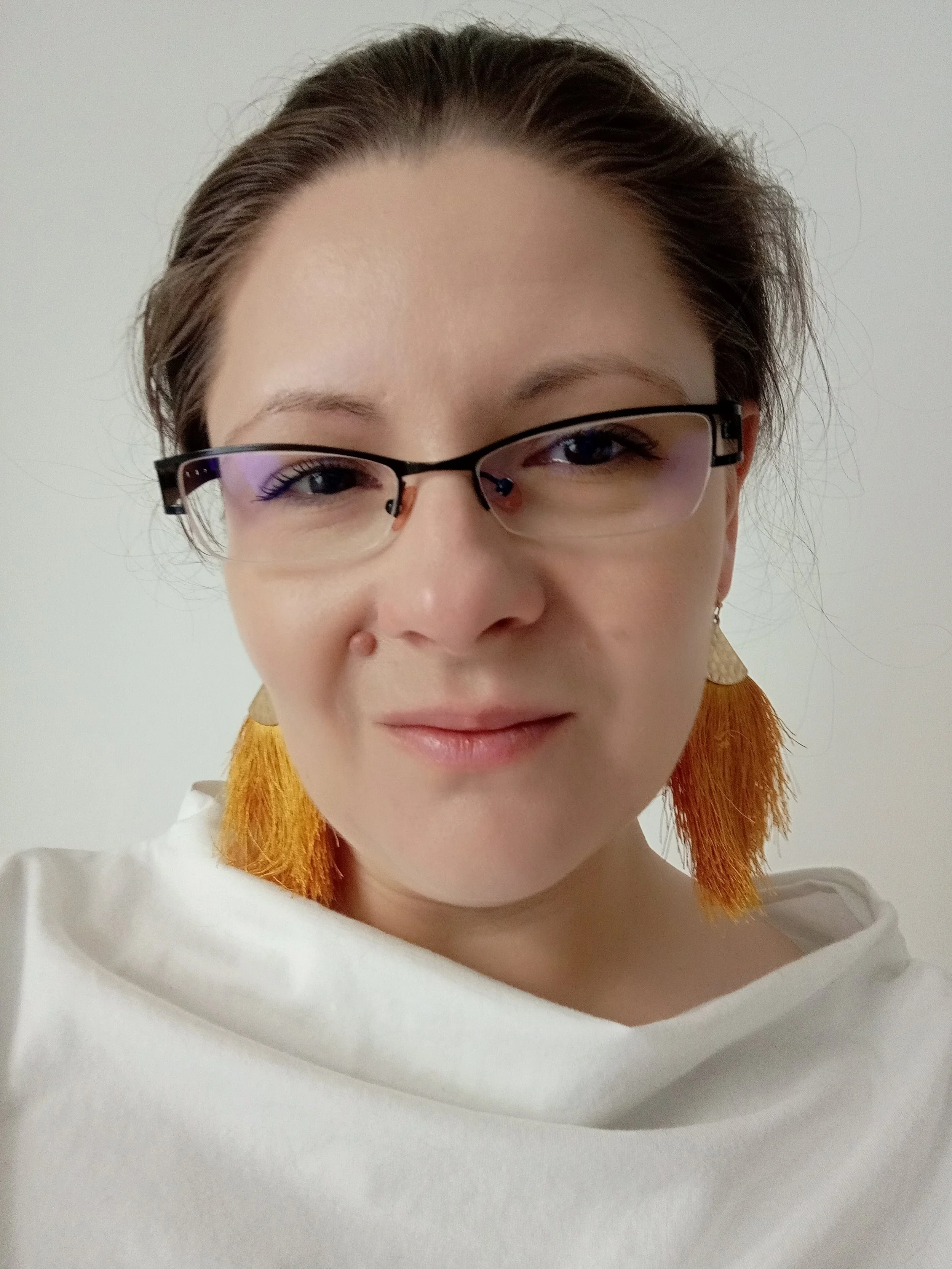 Close-up photo of a woman wearing glasses with purple-tinted lenses and large orange tassel earrings, smiling slightly against a plain white background.