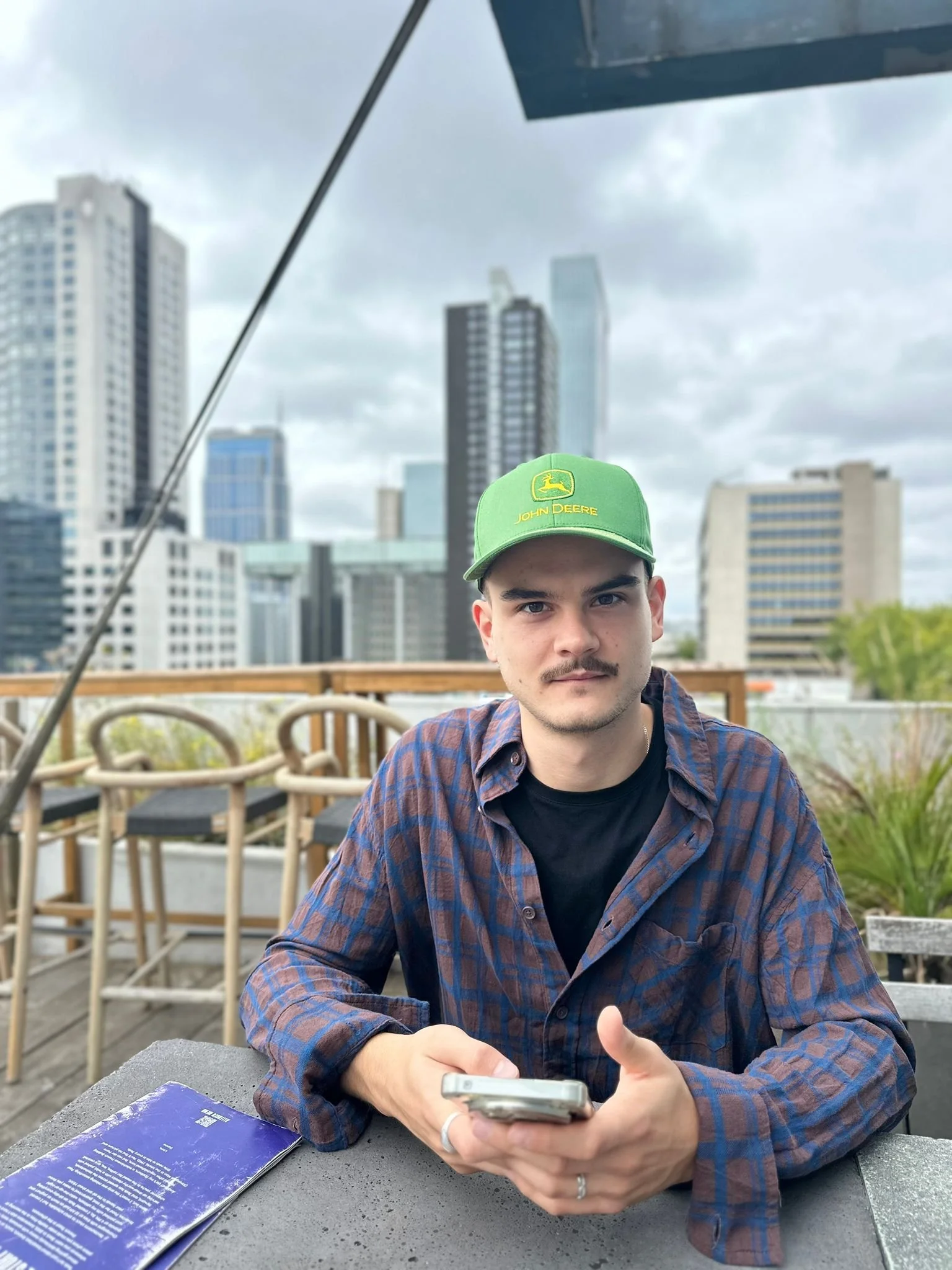 Man sitting at an outdoor table on a rooftop terrace with a city skyline in the background, wearing a green John Deere cap and a blue plaid shirt, holding a phone.