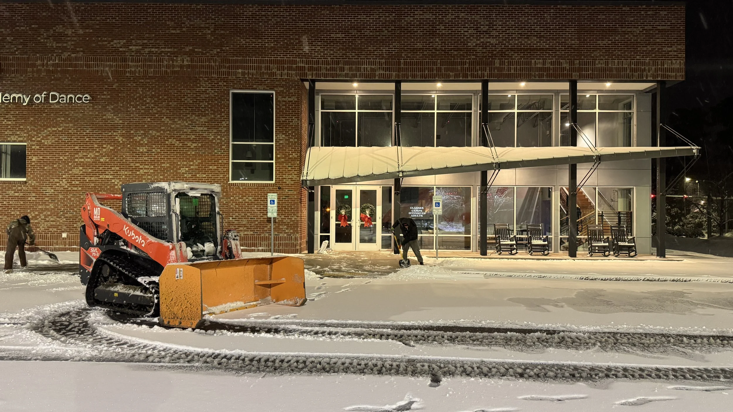 Snow-covered street in front of a brick building with a sign that reads 'Academy of Dance', people shoveling snow, and a small orange snowplow.