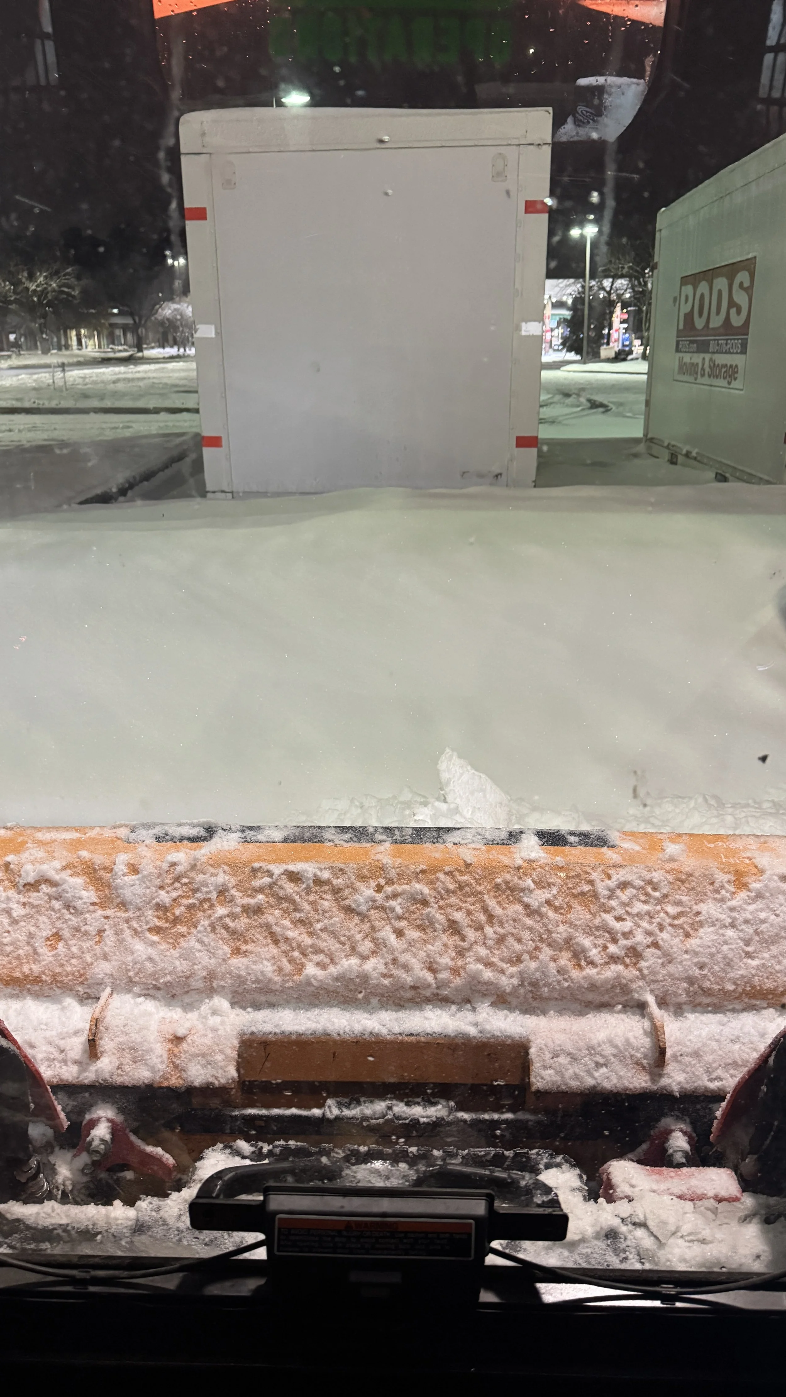 View from inside a snowplow clearing snow at night, with snow-covered parking lot and buildings visible through the windshield. The snowplow's metal blade is pushing snow in front.
