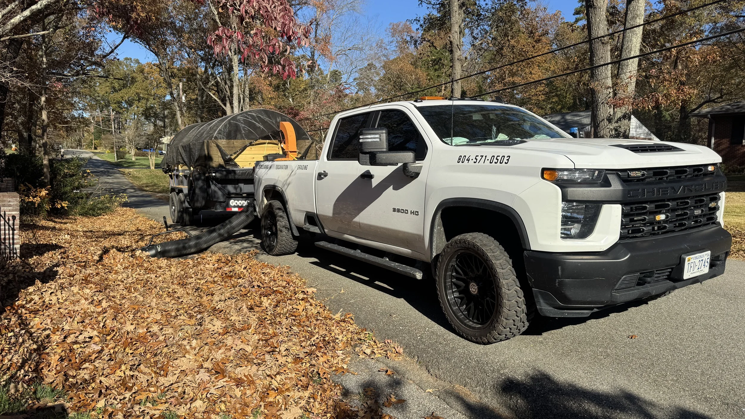 A white Chevrolet truck towing a leaf collection trailer with a leaf blower hose attached, parked on a residential street surrounded by fall foliage.